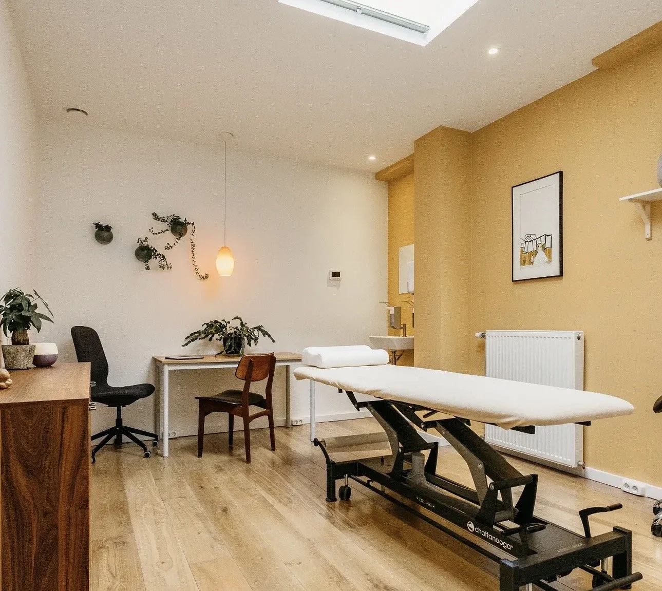 Een medische behandelkamer met een behandeltafel, bureaus, stoelen, planten en muurdecoraties.