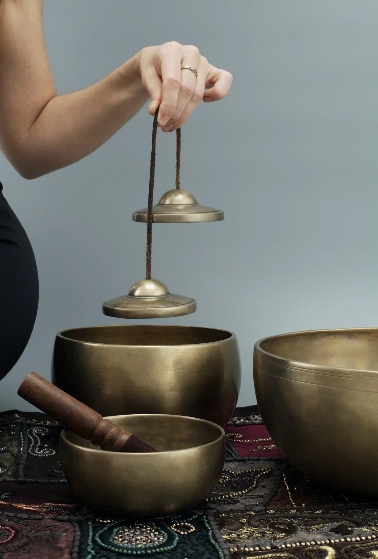 Een hand plaatst een Tibetaanse klankschaal boven een andere, met meerdere klankschalen en sieraden zoals kralen en een houten mallet ernaast op een sierlijk stoffen kleed.