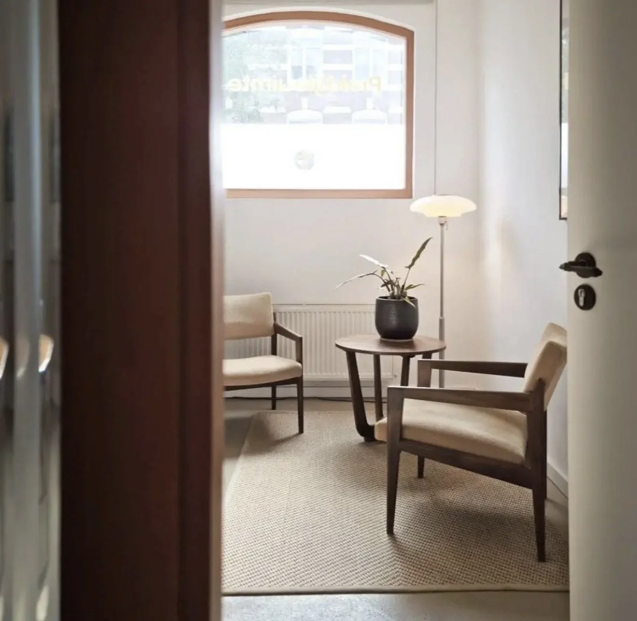 Een lichte kamer met twee beige stoffe stoelen, een ronde houten tafel met een plant in een zwarte pot, een witte lamp, een raam met uitzicht op een gebouw, en een radiator onder het raam.