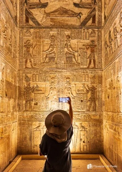 Person wearing a hat taking a selfie with an ancient Egyptian wall of hieroglyphs and carvings.