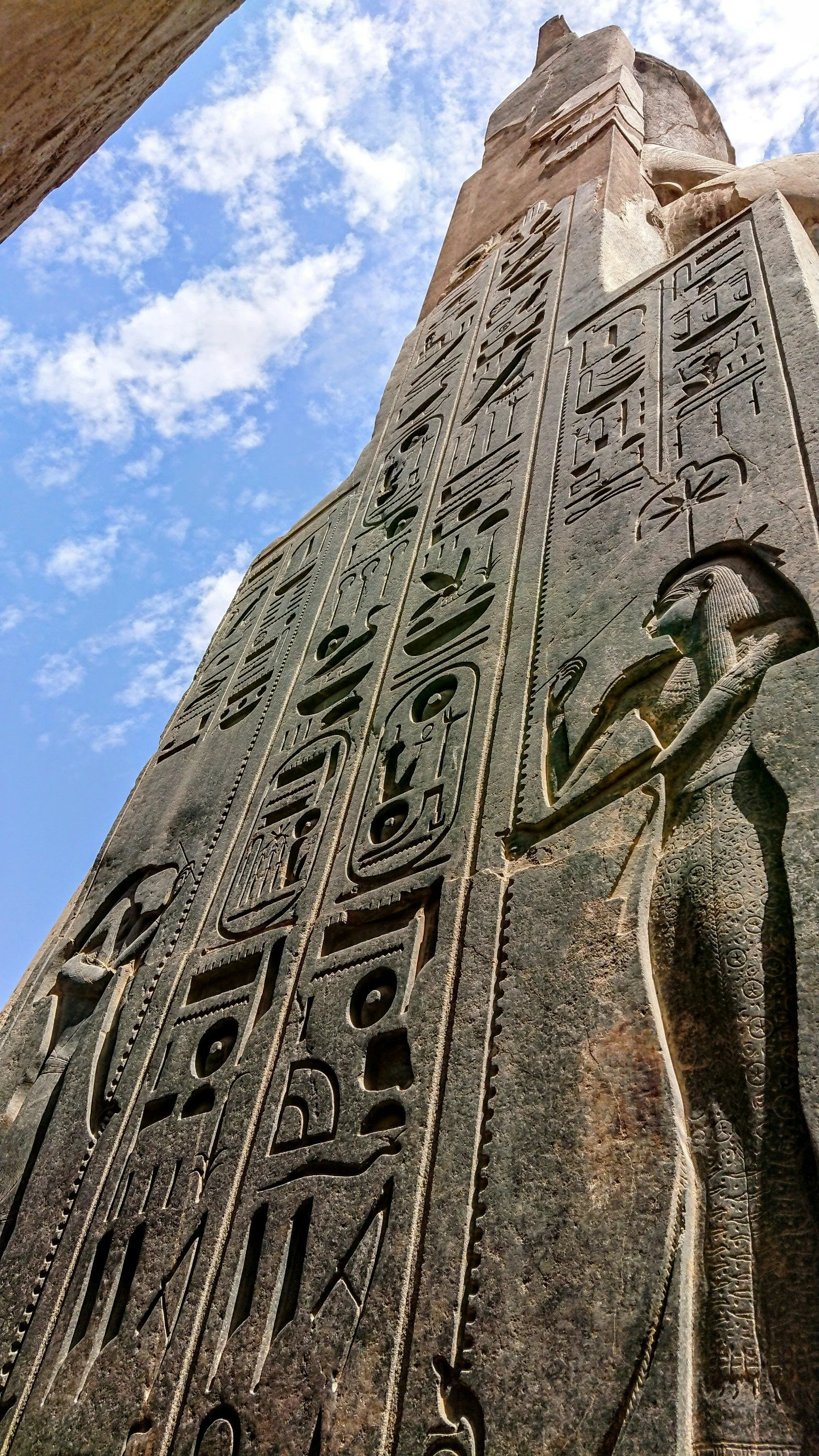 Low angle view of an ancient Egyptian stone monument with hieroglyphics and carvings, set against a partly cloudy sky.