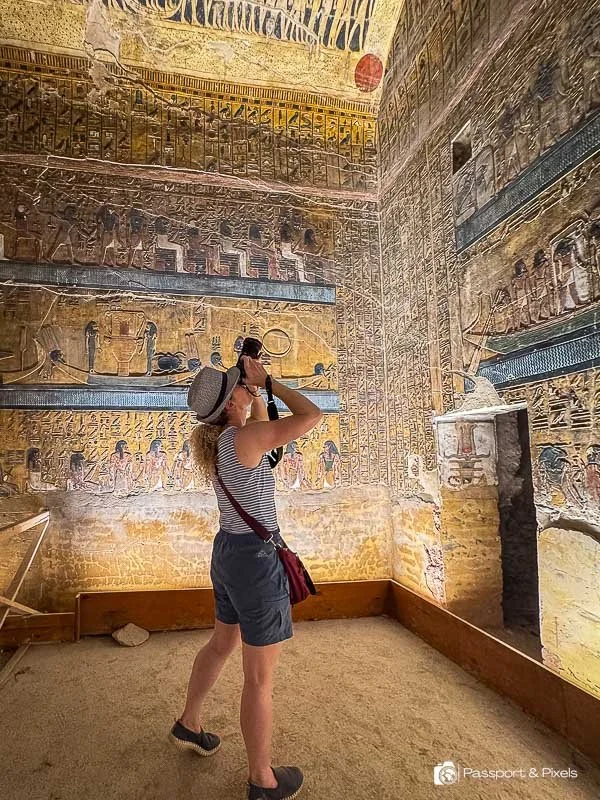 A woman with a backpack and hat taking a photo with her phone in an ancient Egyptian tomb decorated with hieroglyphics and wall paintings.