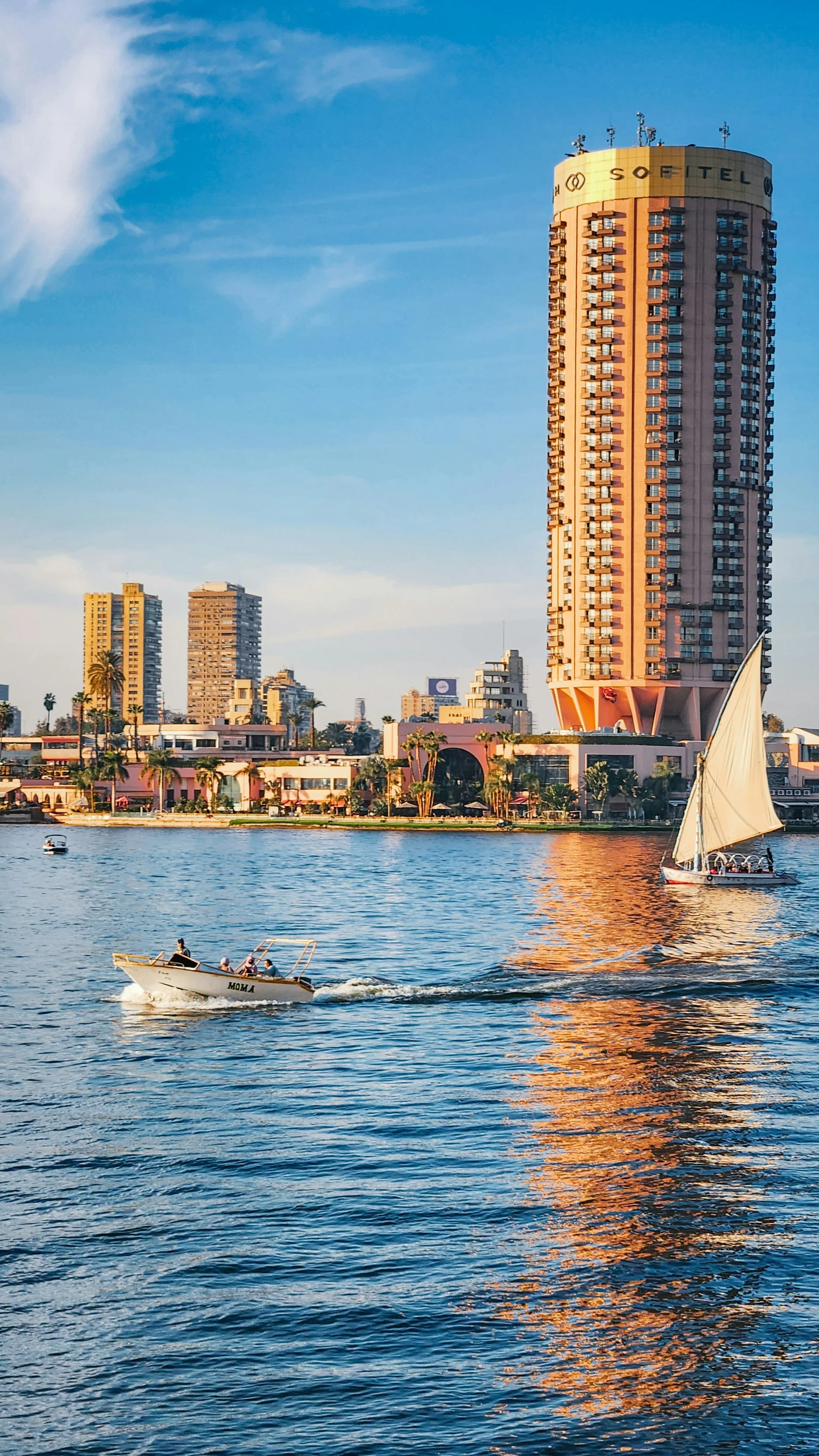 A city skyline with high-rise buildings along a body of water, featuring boats and a sailboat, during sunset with clear blue sky.
