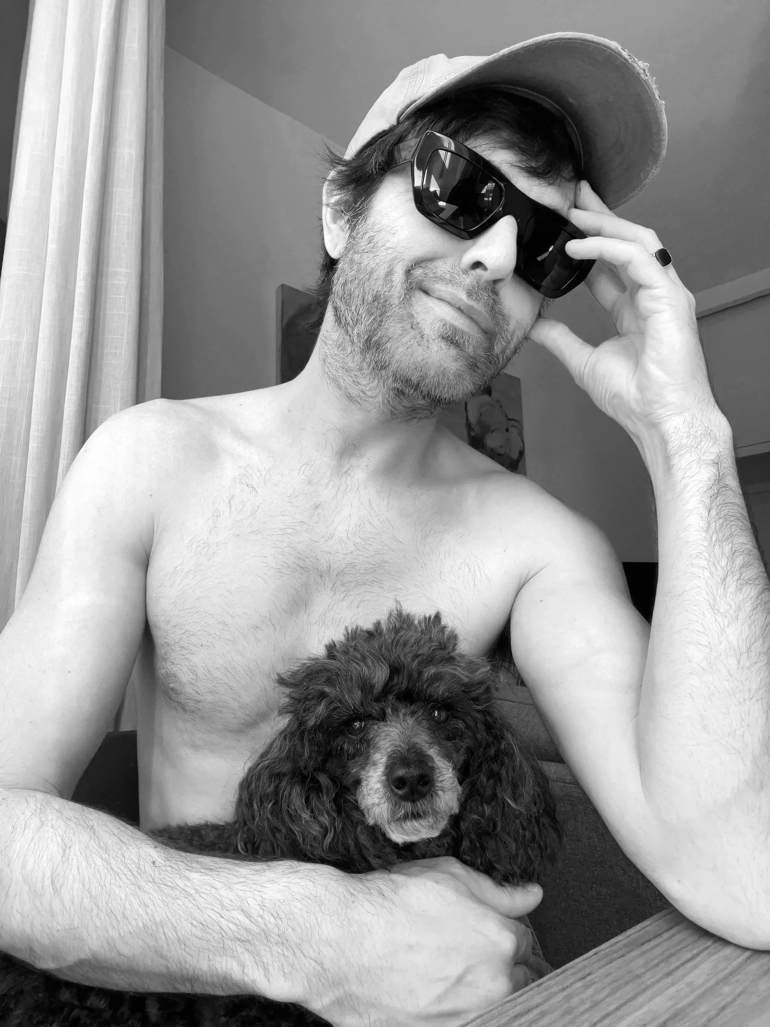 A shirtless man with sunglasses, a cap, and a ring smiles while holding a puppy, sitting indoors near a window with curtains.
