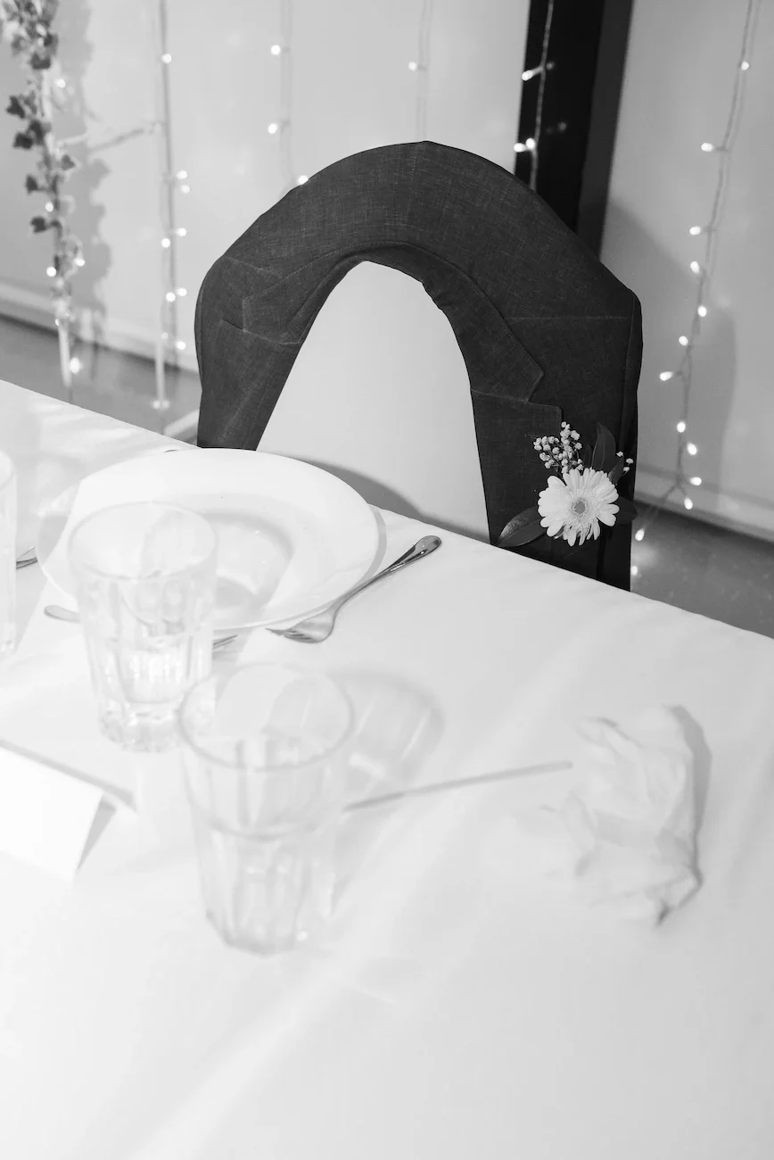 A decorated dining table with empty glasses, a plate, and a fork, with a black chair featuring a flower decoration, and string lights in the background.