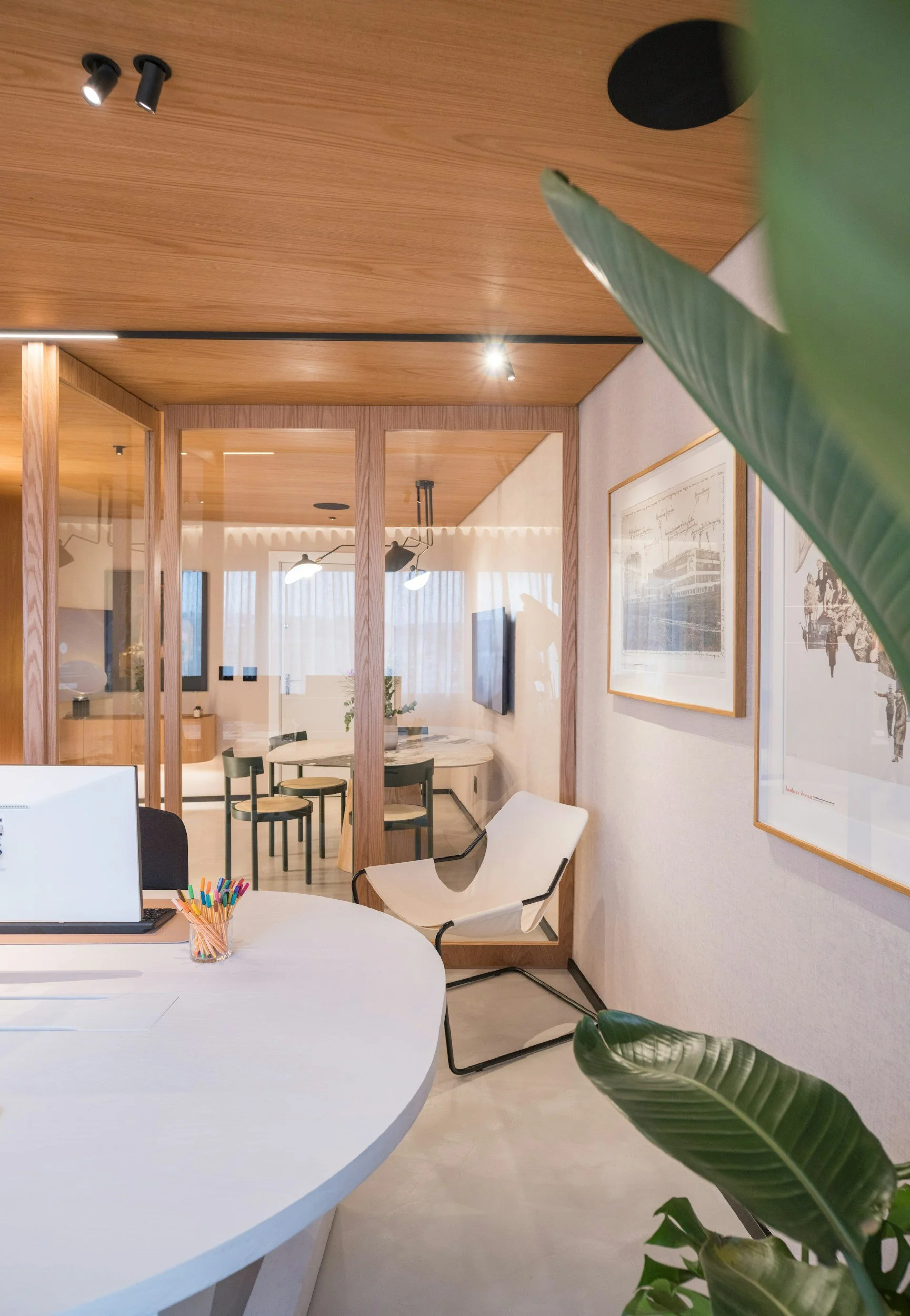 Modern office with a white round desk, a laptop, a container of colored pens, and a white ergonomic chair. Behind a wooden partition, there is a conference area with a round table and black chairs. The office has wooden ceiling panels, framed artwork on the walls, and large windows with sheer curtains, allowing natural light to fill the space.