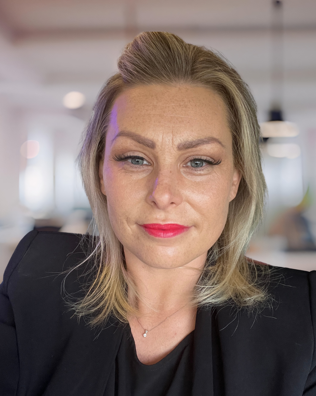 A photo of Gill.  She has blonde, shoulder-length hair, wearing red lipstick, a black top, and a silver necklace, in an office setting.