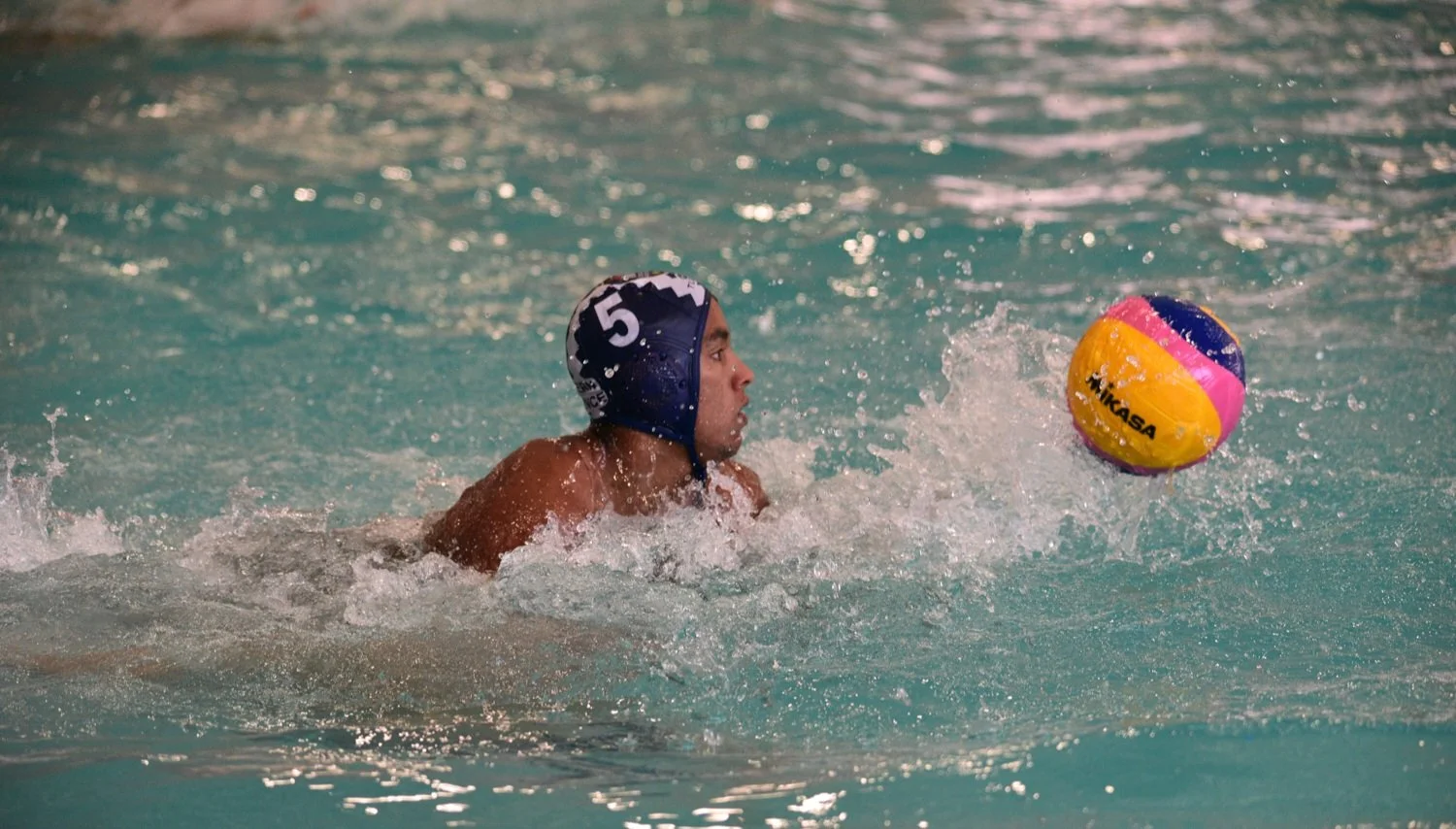 A water polo player wearing a blue cap with the number 5 is swimming in a pool and reaching for a yellow, pink, and purple Mikasa water polo ball.
