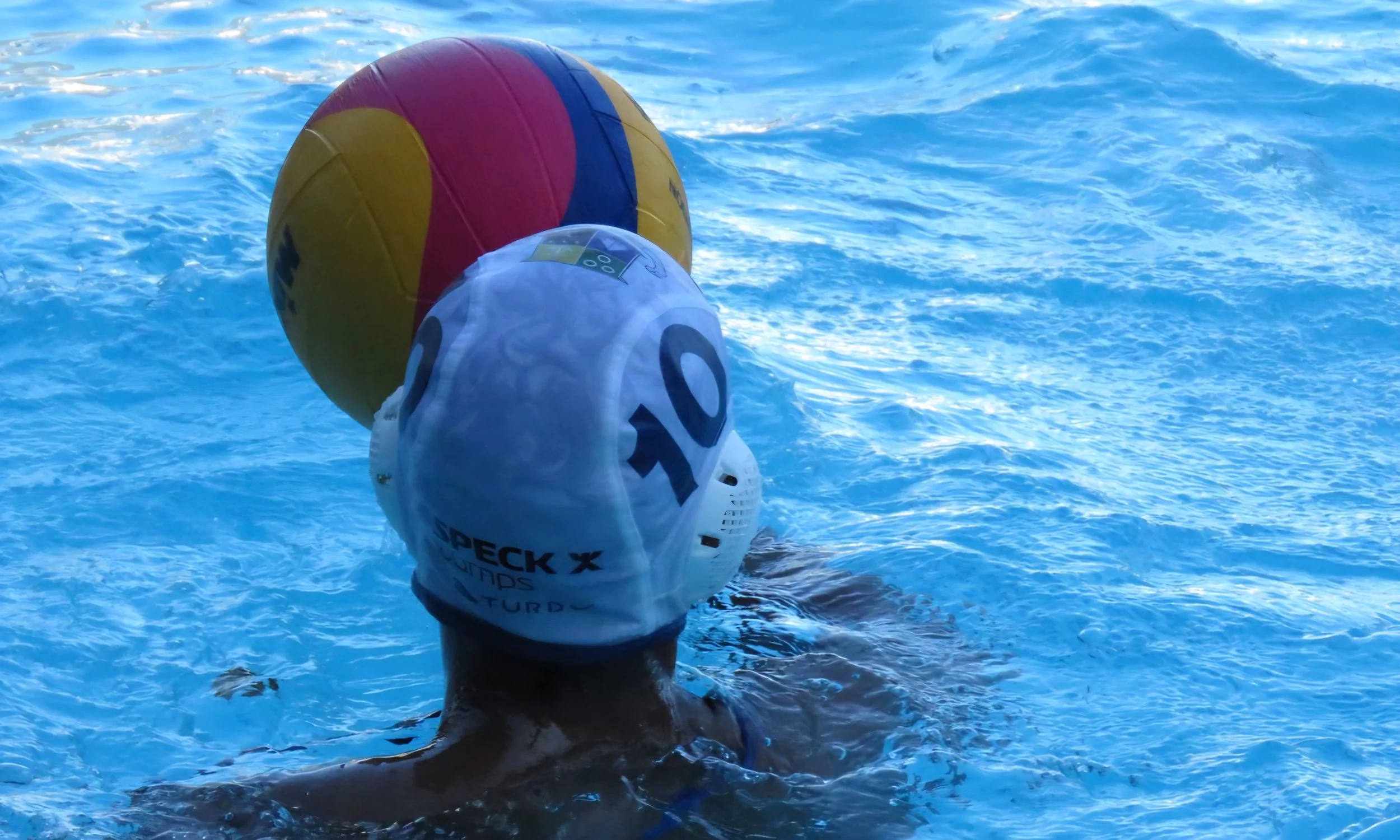 A swimmer in a swimming cap with text on it, holding their breath underwater, with a large colorful beach ball floating nearby in a swimming pool with bright blue water.