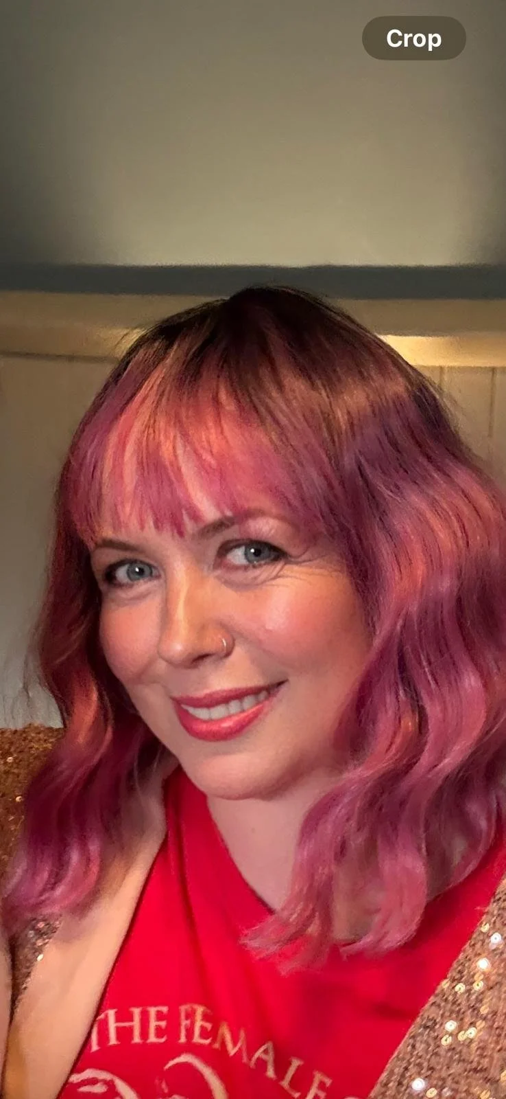 A woman with wavy, pinkish-red hair, blue eyes, and a nose piercing, smiling and wearing a red top with some gold sequined clothing.