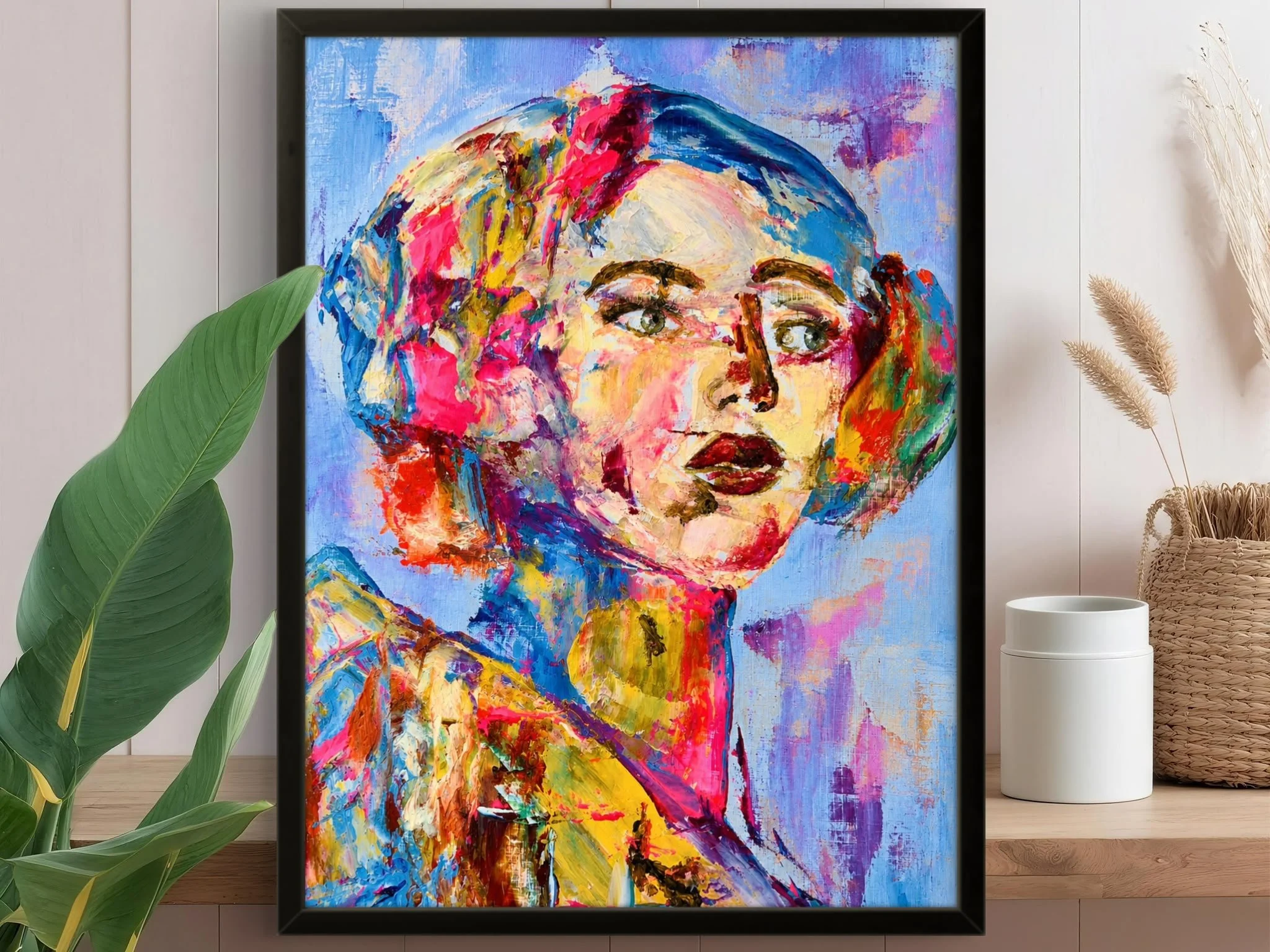 A colorful abstract painting of a woman's face, with bright splashes of pink, yellow, blue, and purple, framed in black and displayed on a white wall.