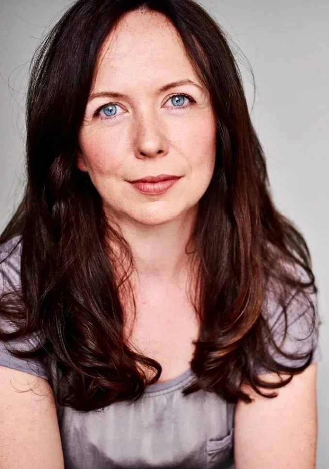 A woman with long, wavy brown hair and blue eyes, wearing a light purple top, smiling softly against a plain gray background.