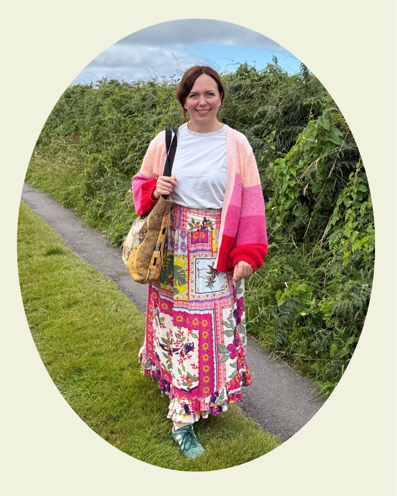 Smiling woman standing outdoors on a grassy path, wearing a colorful embroidered skirt, white t-shirt, pink and red cardigan, teal sneakers, and carrying a shoulder bag with a patchwork design, with green bushes and a cloudy sky in the background.