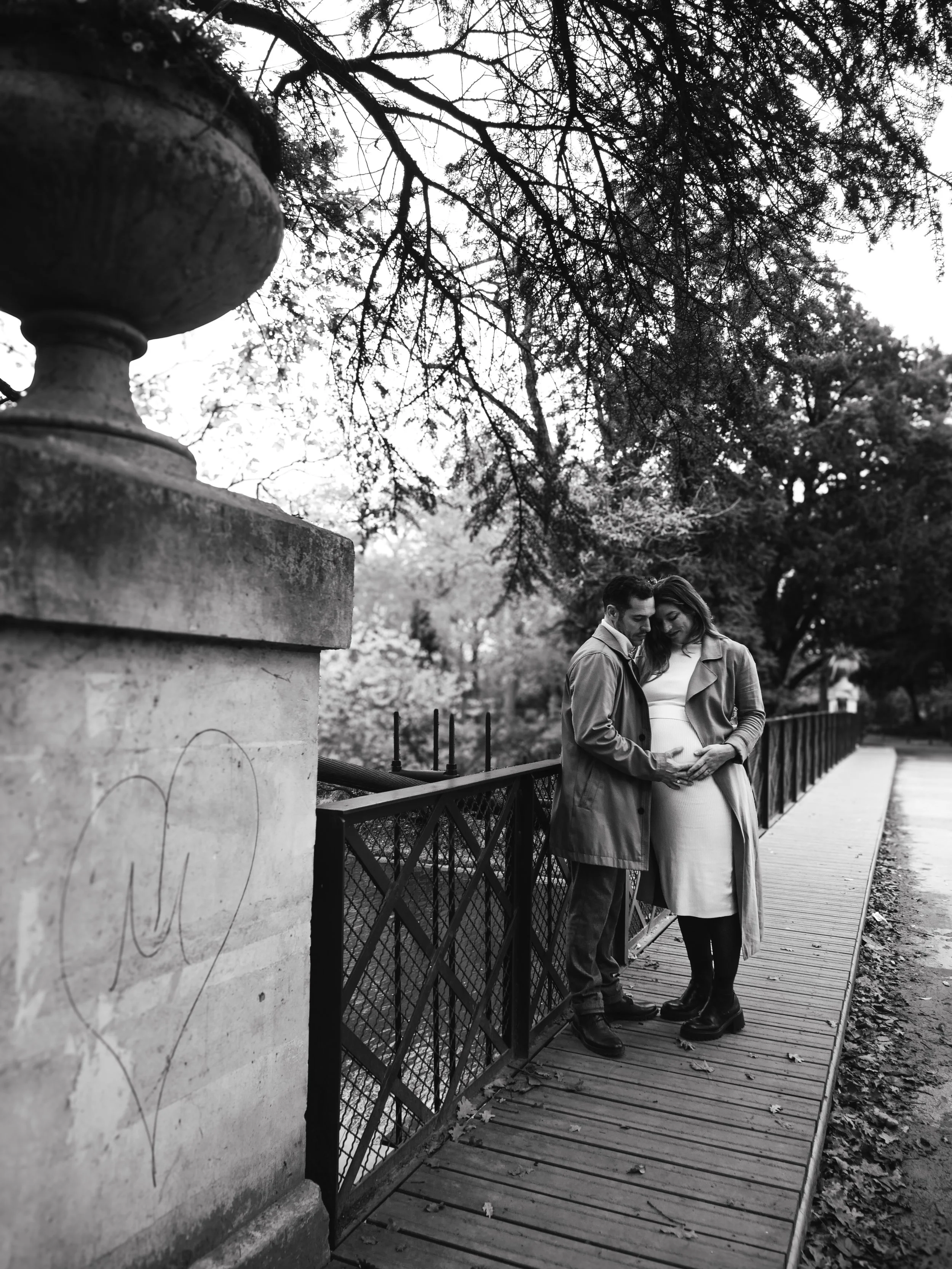 Un couple debout côte à côte sur un pont en bois, se tenant la main et se regardant tendrement. La femme semble enceinte. L'arrière-plan montre des arbres feuillus et un ciel clair.