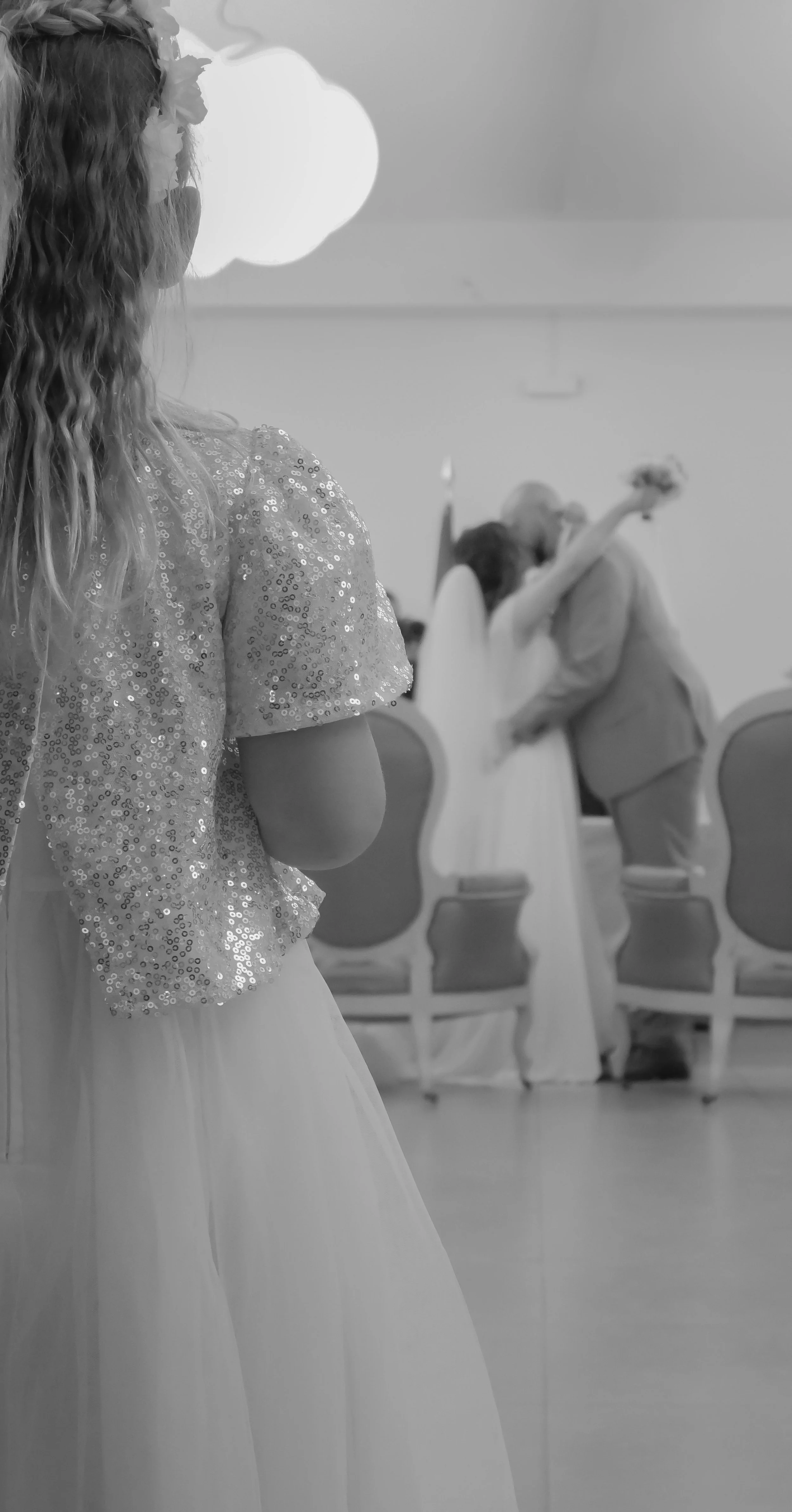 Une jeune fille en robe de bal avec un haut en sequins, vue de dos, observant un bal où un couple danse.