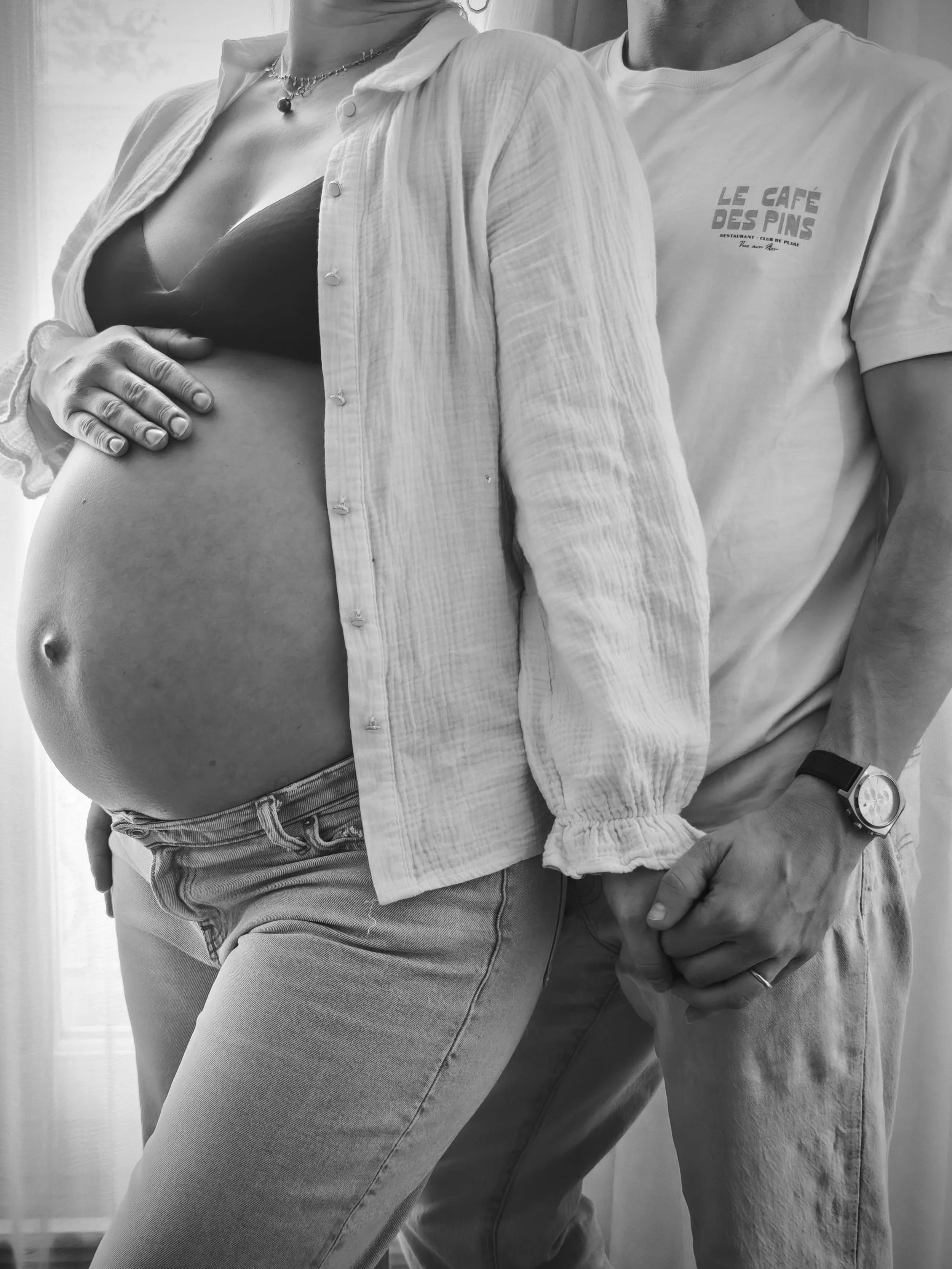 Une femme enceinte en crop top et cardigan, avec un collier, et un homme à côté d'elle qui tient sa main. La photo est en noir et blanc.