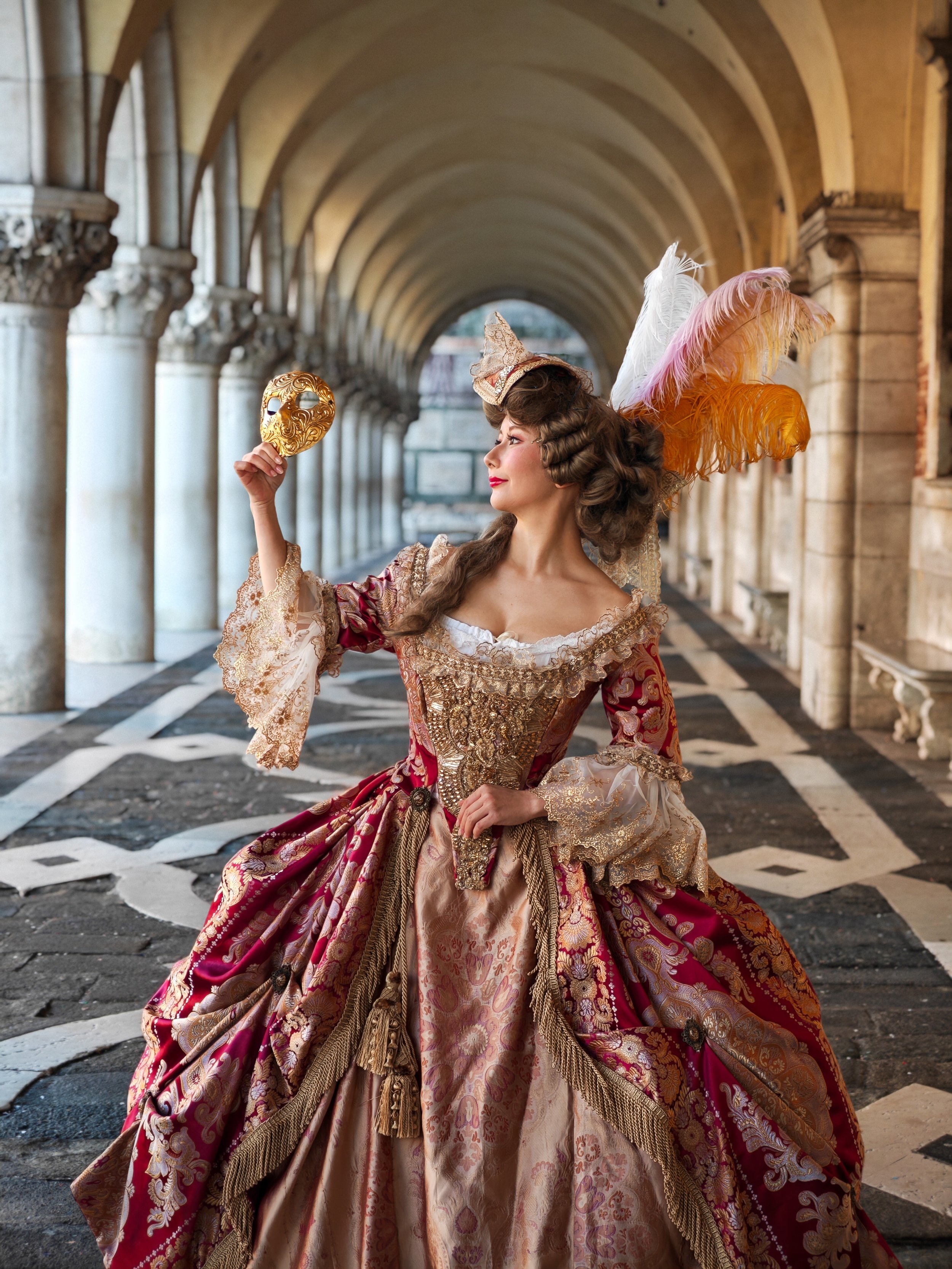 Une femme habillée d'un costume historique avec un masque doré, un chapeau orné de plumes, dans un couloir arcades en pierre, portant une robe richement décorée et tenant un masque doré.