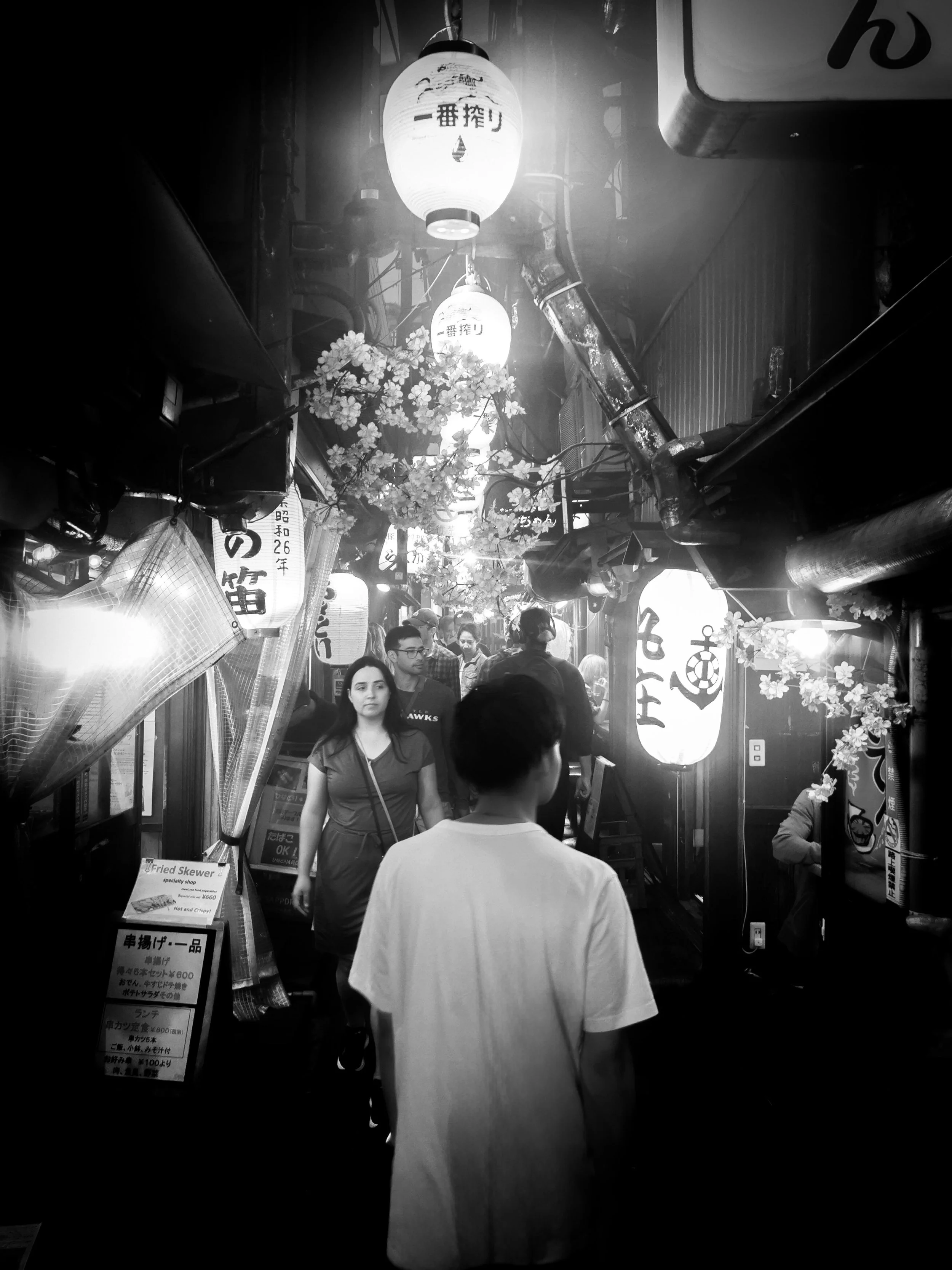 Une vue de rue animée à l'entrée d'un restaurant japonais avec lanternes traditionnelles éclairantes, des personnes discutant et une décoration florale.