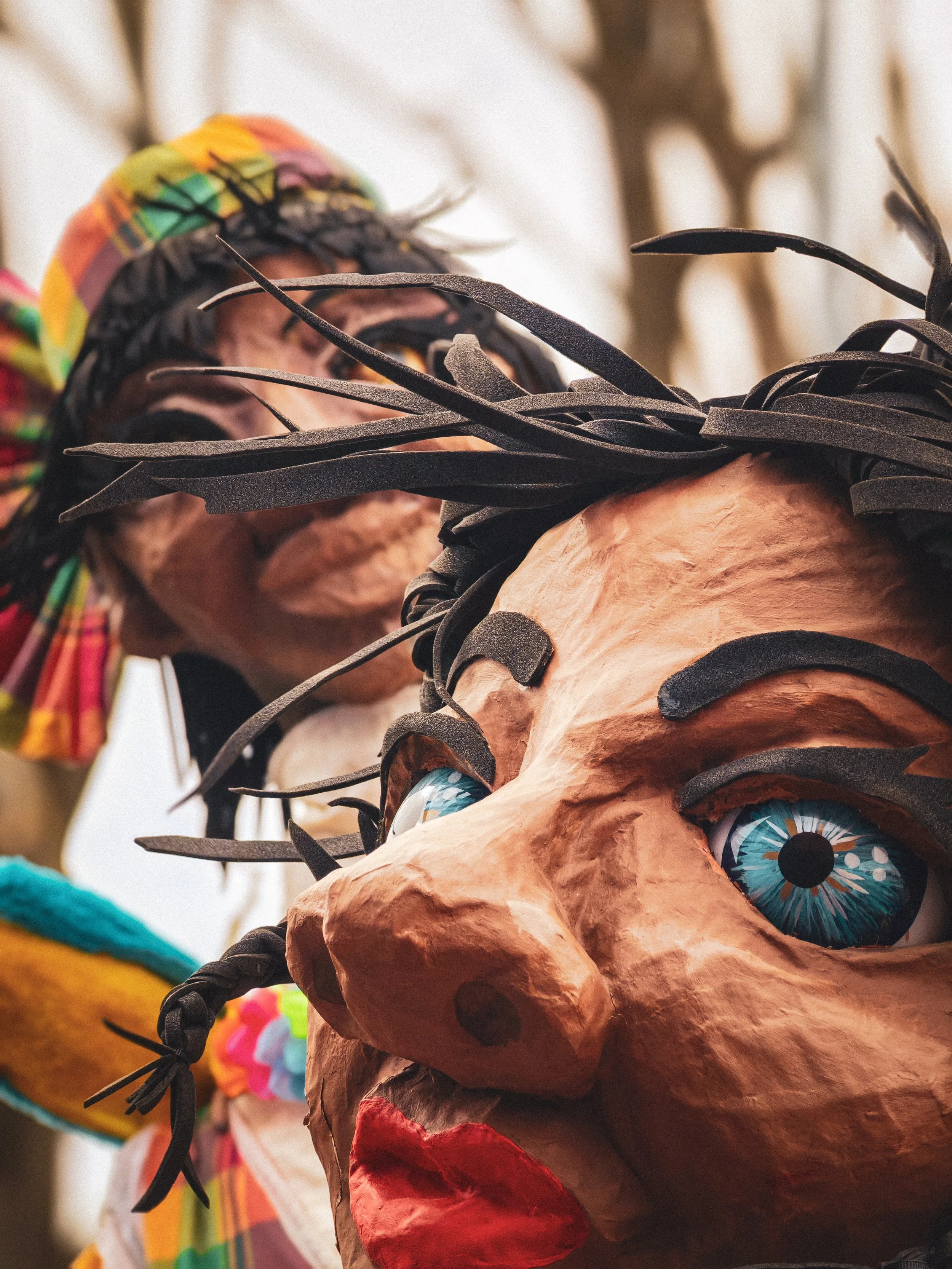 Close-up d'une tête de masque en papier mâché représentant un personnage avec des yeux bleus, des cheveux noirs en broussaille, un nez proéminent et une bouche rouge. Le masque a une apparence colorée et expressive.