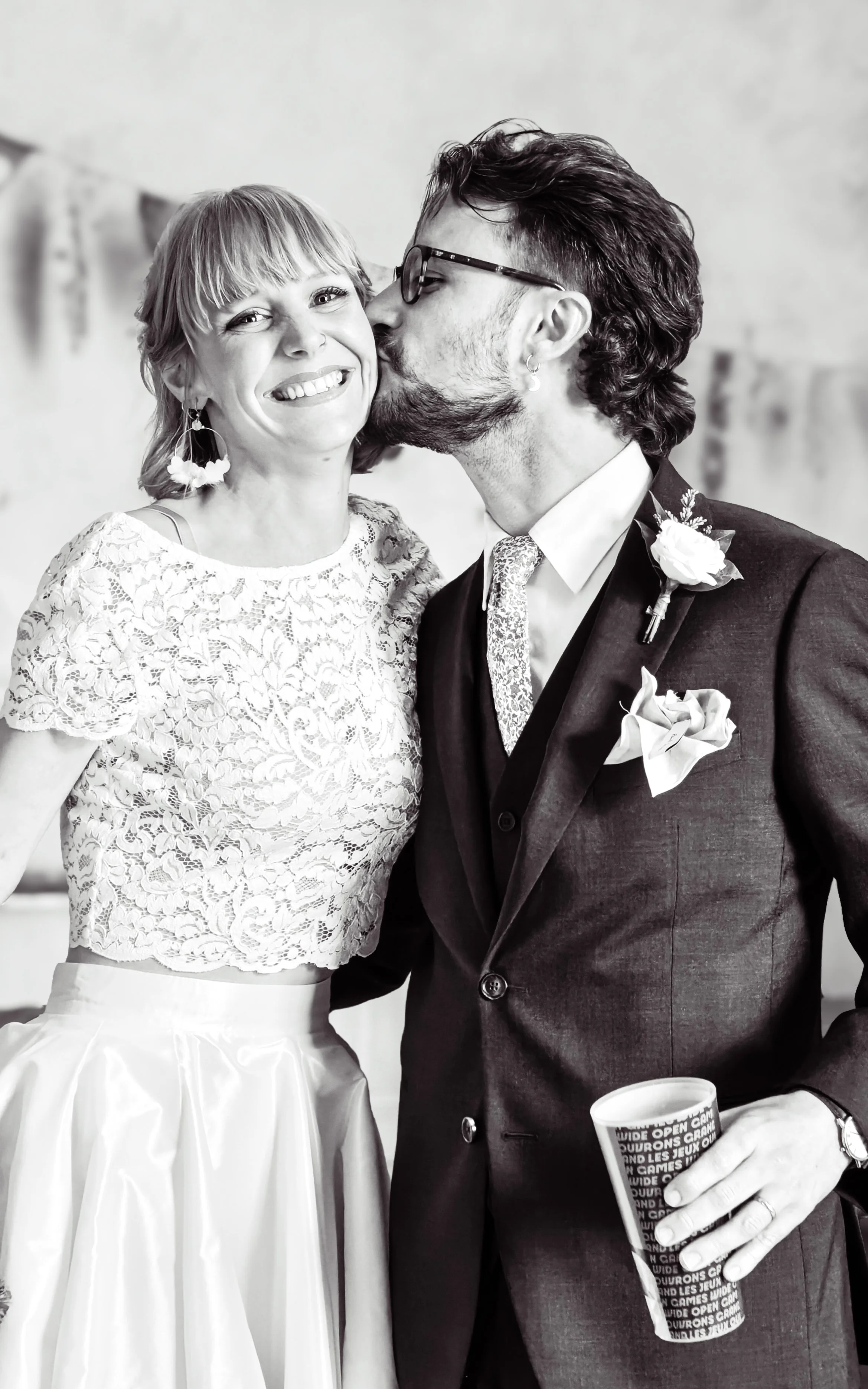 Un couple lors d'une cérémonie, la femme souriante porte une robe en dentelle avec un crop top et une jupe longue, l'homme portant un costume sombre avec une boutonnière et tenant une tasse racontant une blague humoristique.