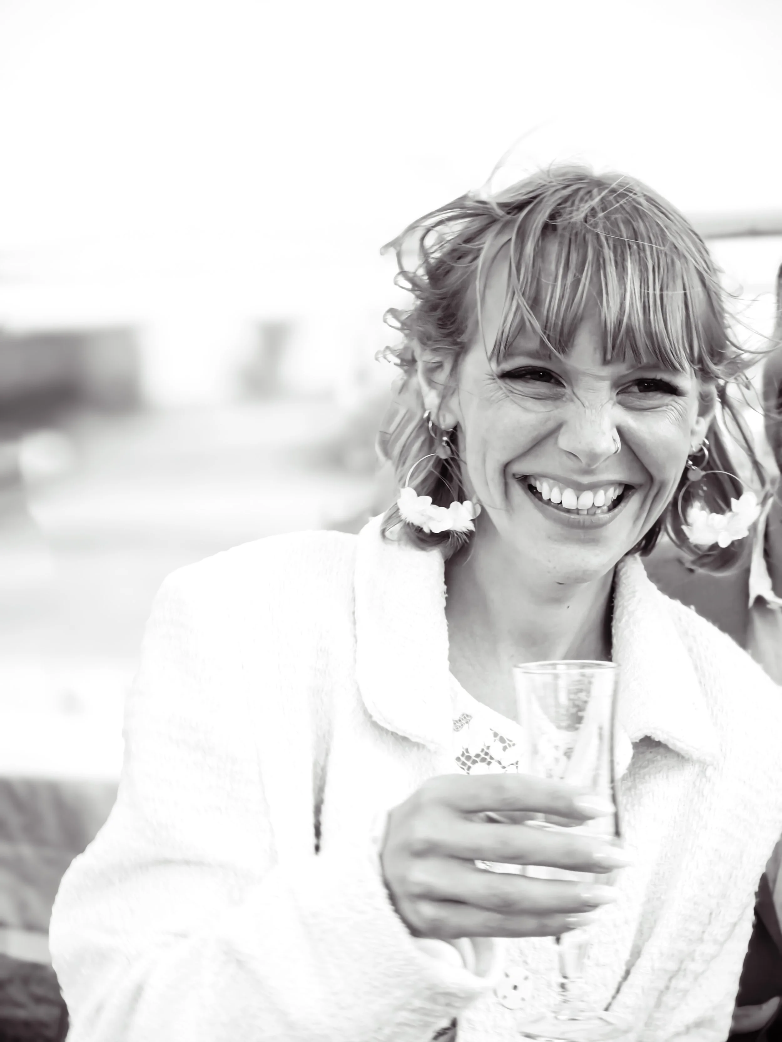 Une femme souriante tenant un verre de boisson, portant des boucles d'oreilles avec des fleurs, en extérieur, en noir et blanc.
