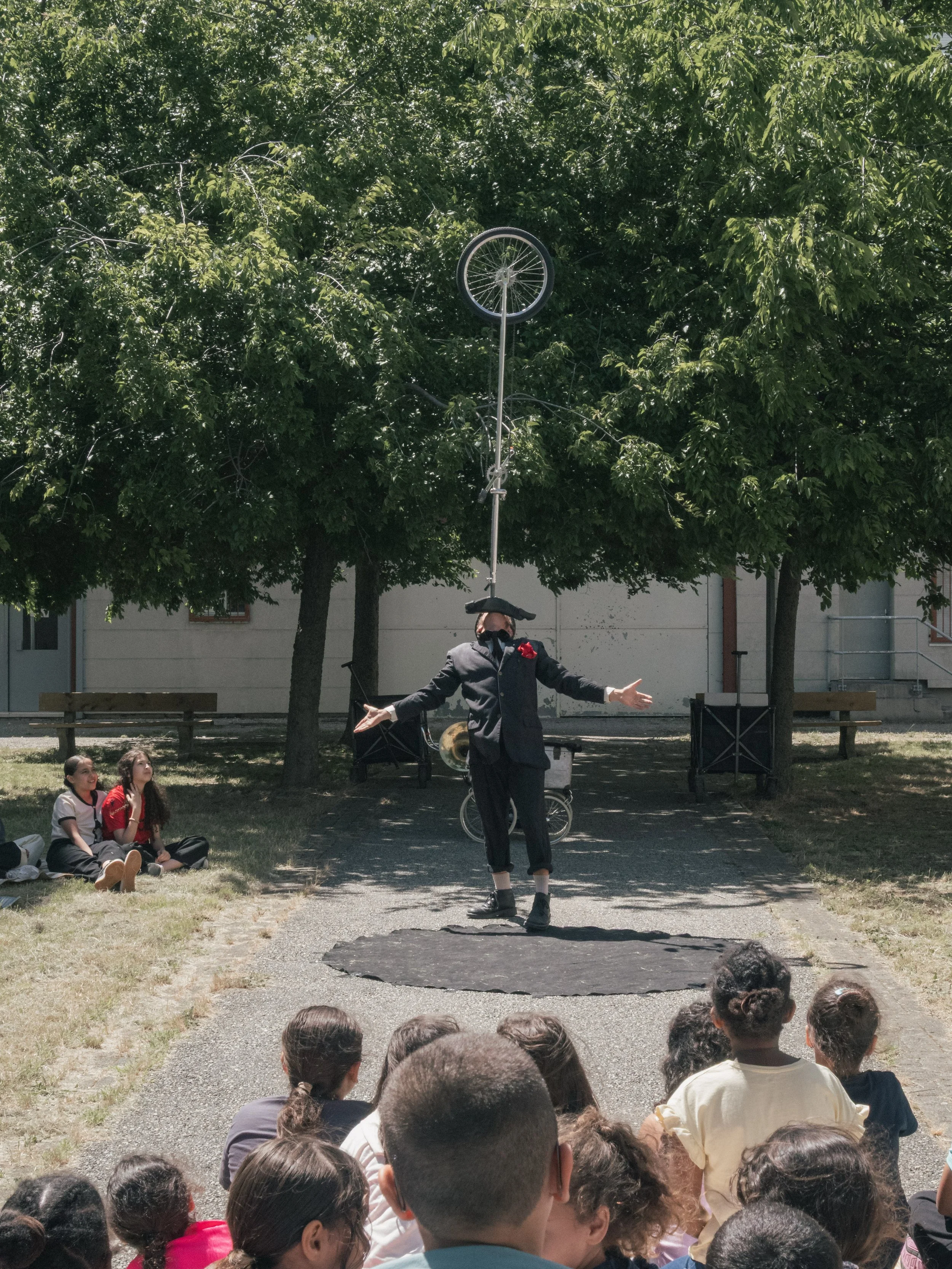 Un jongleur habillé en noir avec un chapeau noir et une fleur rouge effectue un numéro de jonglage avec une roue de vélo suspendue en haut d'une perche, sous un arbre, avec un public d'enfants assis sur l'herbe.