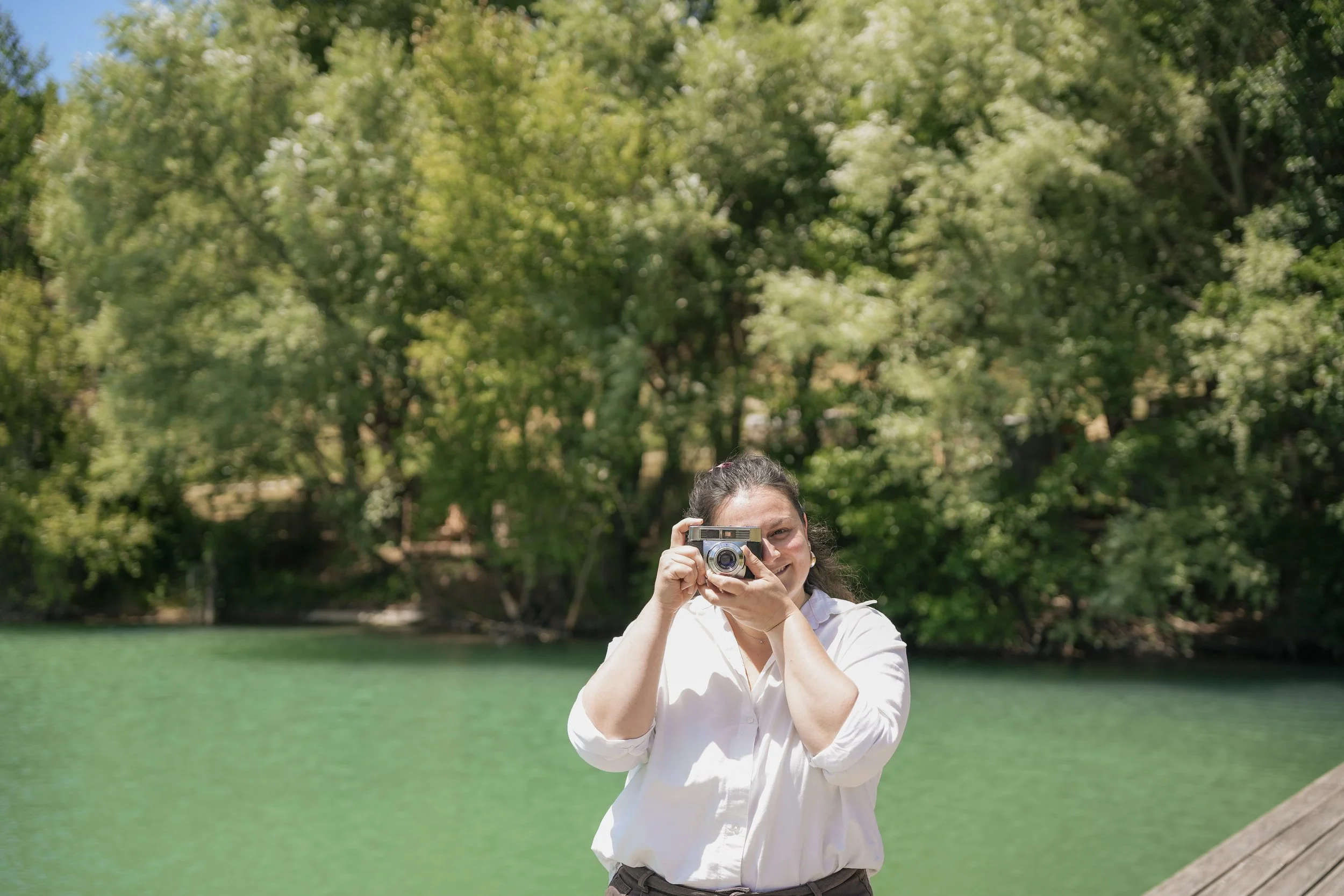 Une femme souriante tenant un appareil photo, en extérieur près d'un étang avec des arbres verts en arrière-plan.