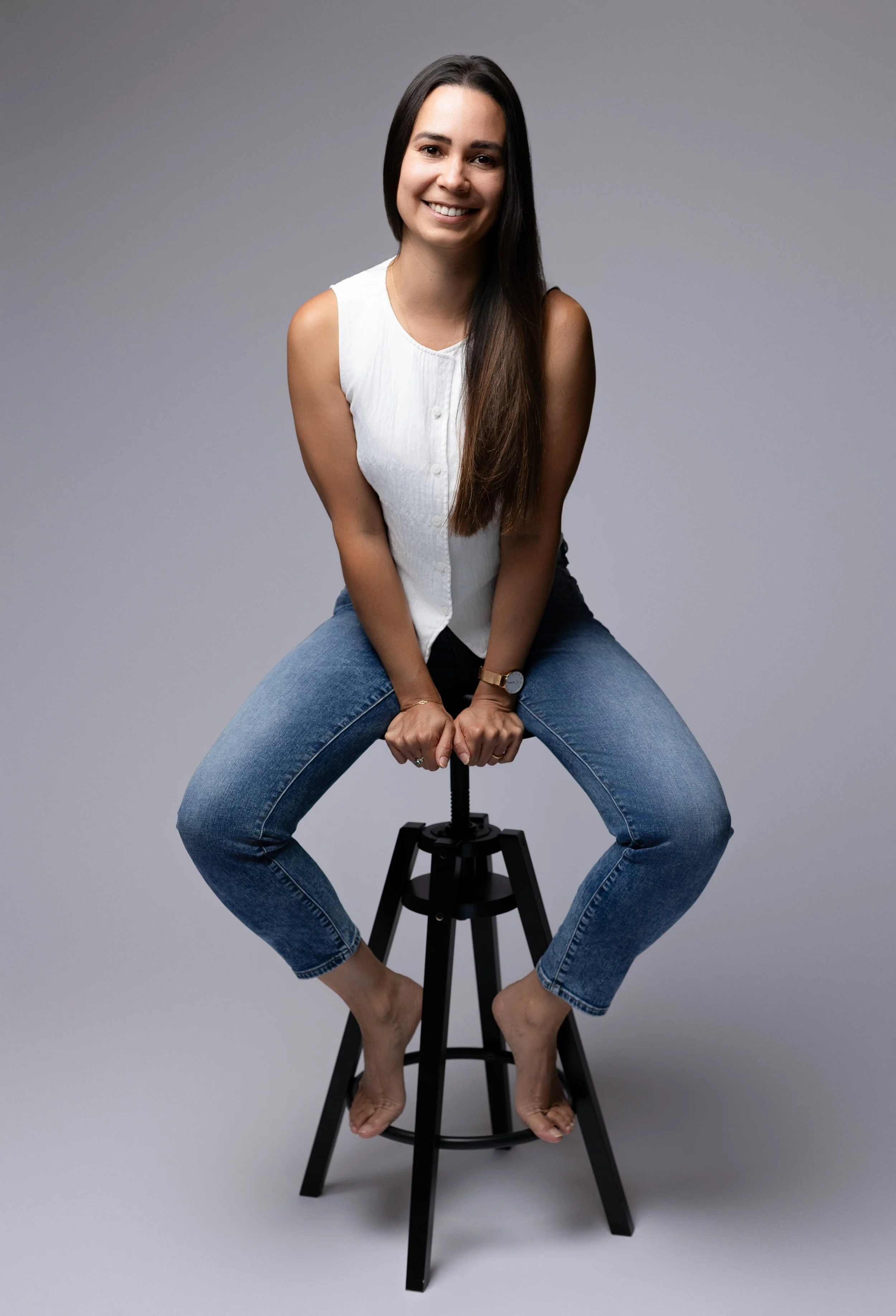 Une femme souriante assise sur un tabouret noir, avec les jambes écartées et pieds nus, portant une blouse blanche et un jean bleu, contre un fond gris.