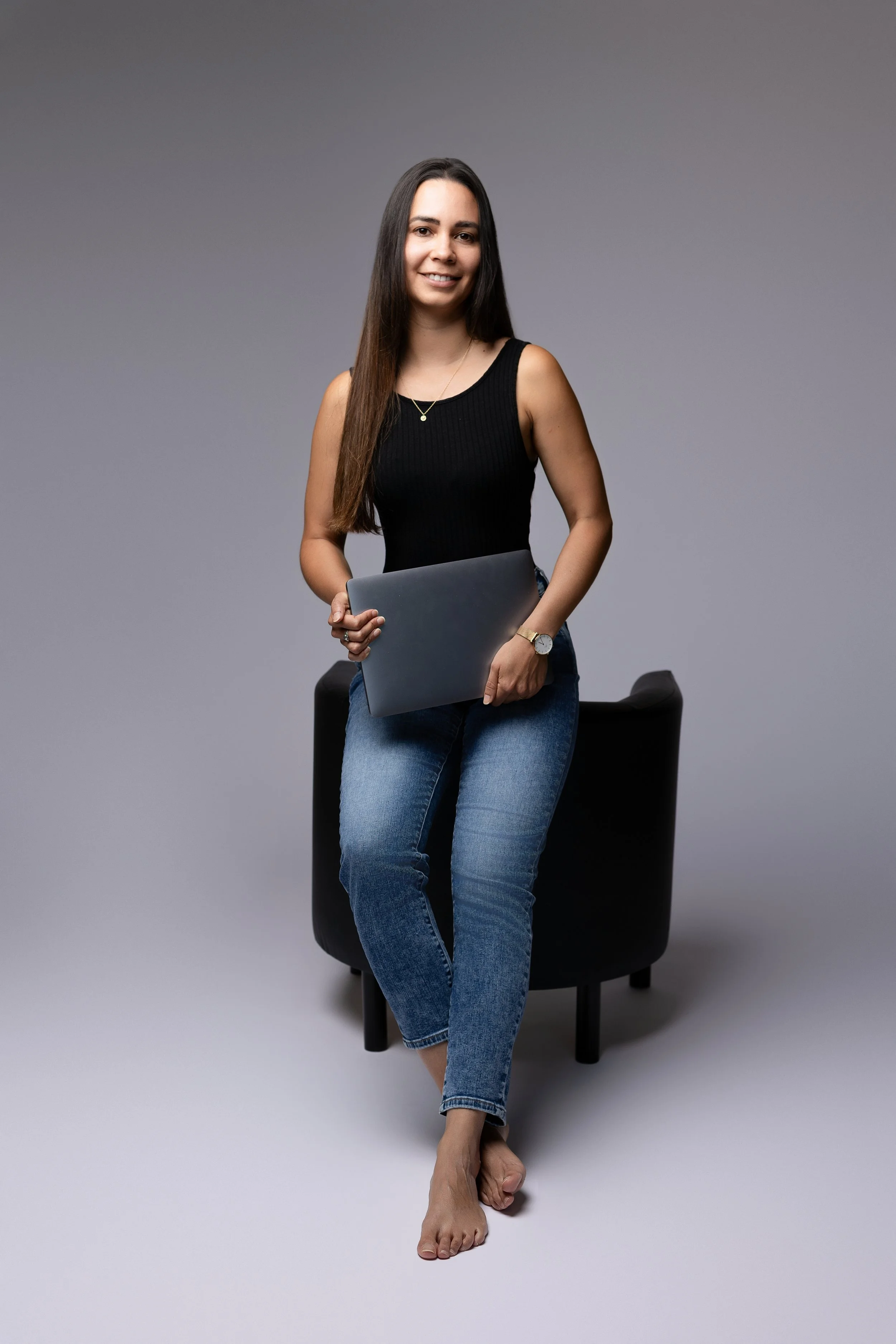 Une femme jeune avec des cheveux longs, portant une chemise sans manches noire, tenant un ordinateur portable, assise sur une chaise noire en face d'un fond gris, avec une montre et une chaîne en or comme bijoux, et souriante.