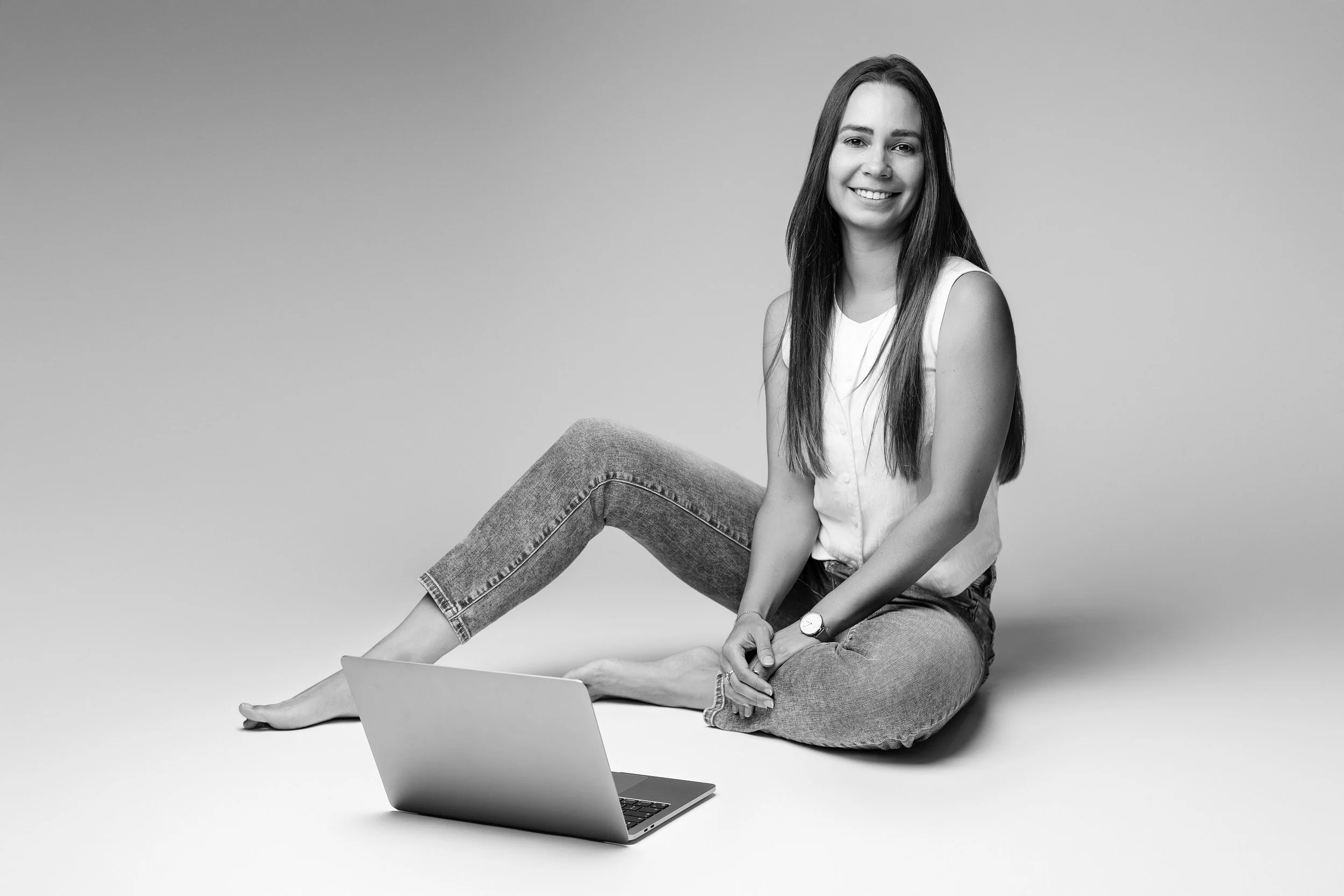 Jeune femme souriante assise par terre avec un ordinateur portable, regardant vers l'avant, vêtue d'un débardeur blanc et de jeans, fond gris