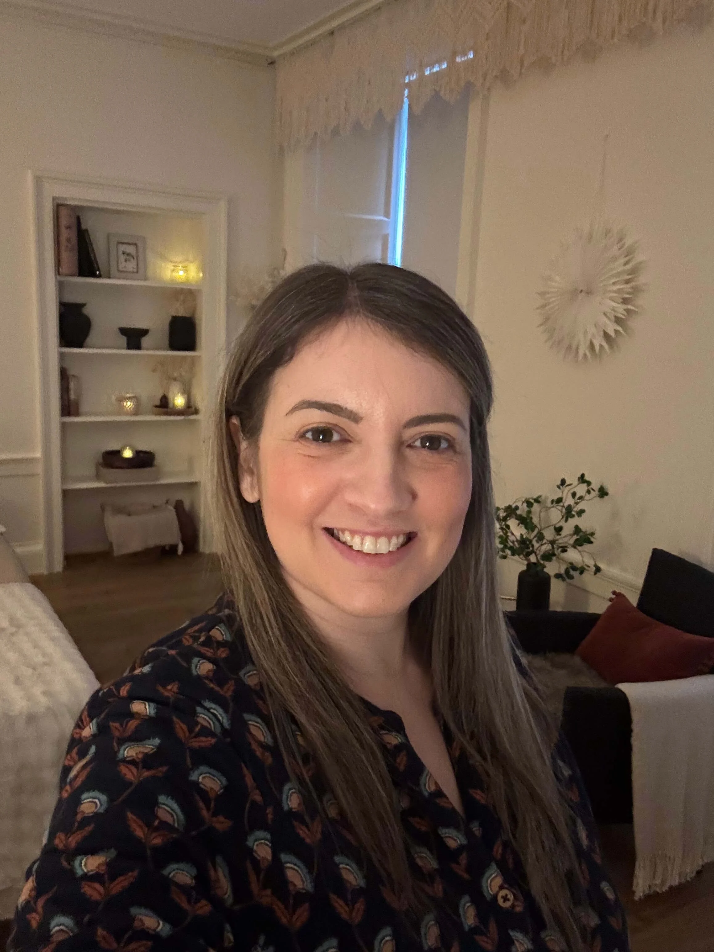 A woman smiling indoors, with a Reiki room background featuring a bookshelf with decorative items, a window with curtains, and some wall art