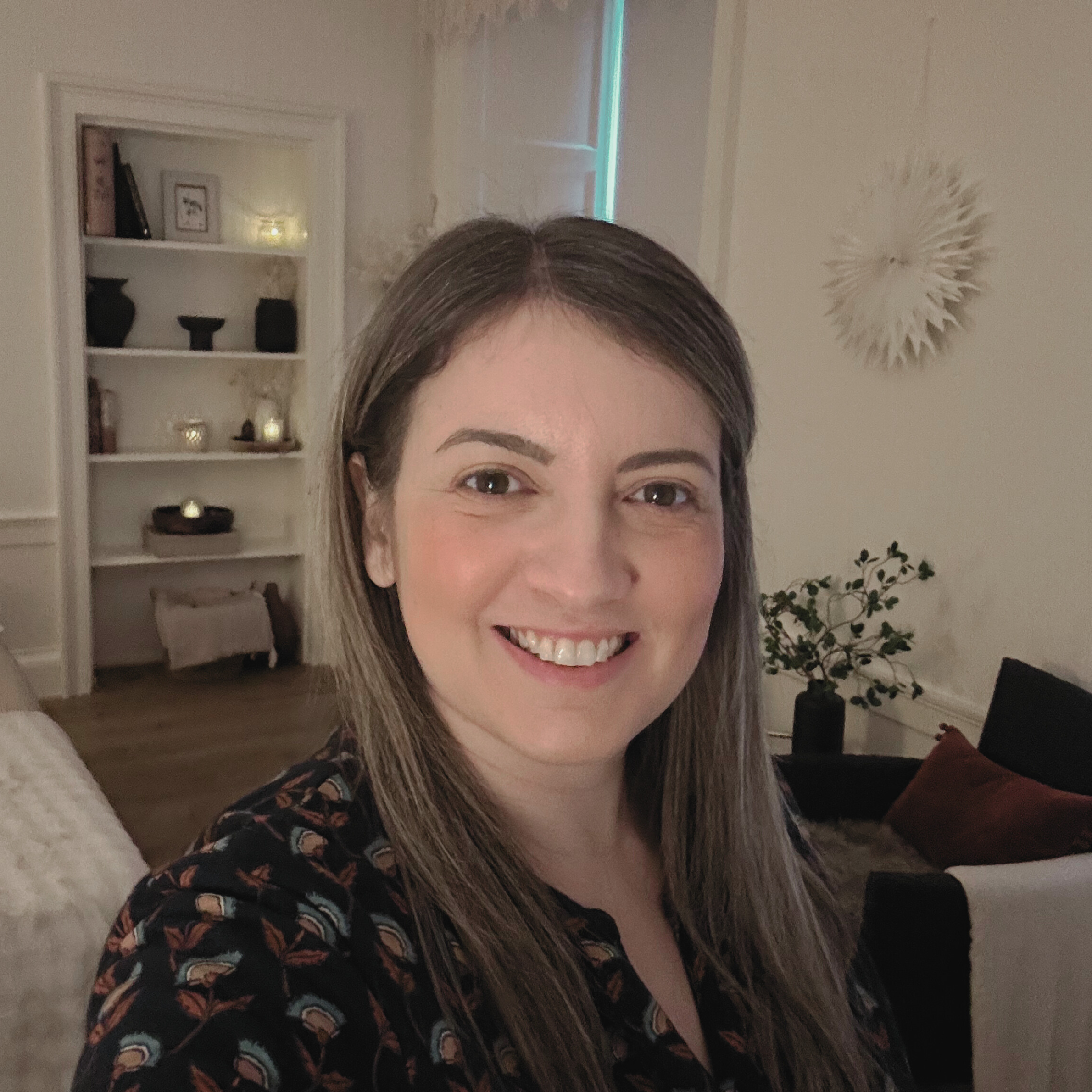 A woman with brown hair taking a selfie in a room with white walls and a bookshelf in the background.