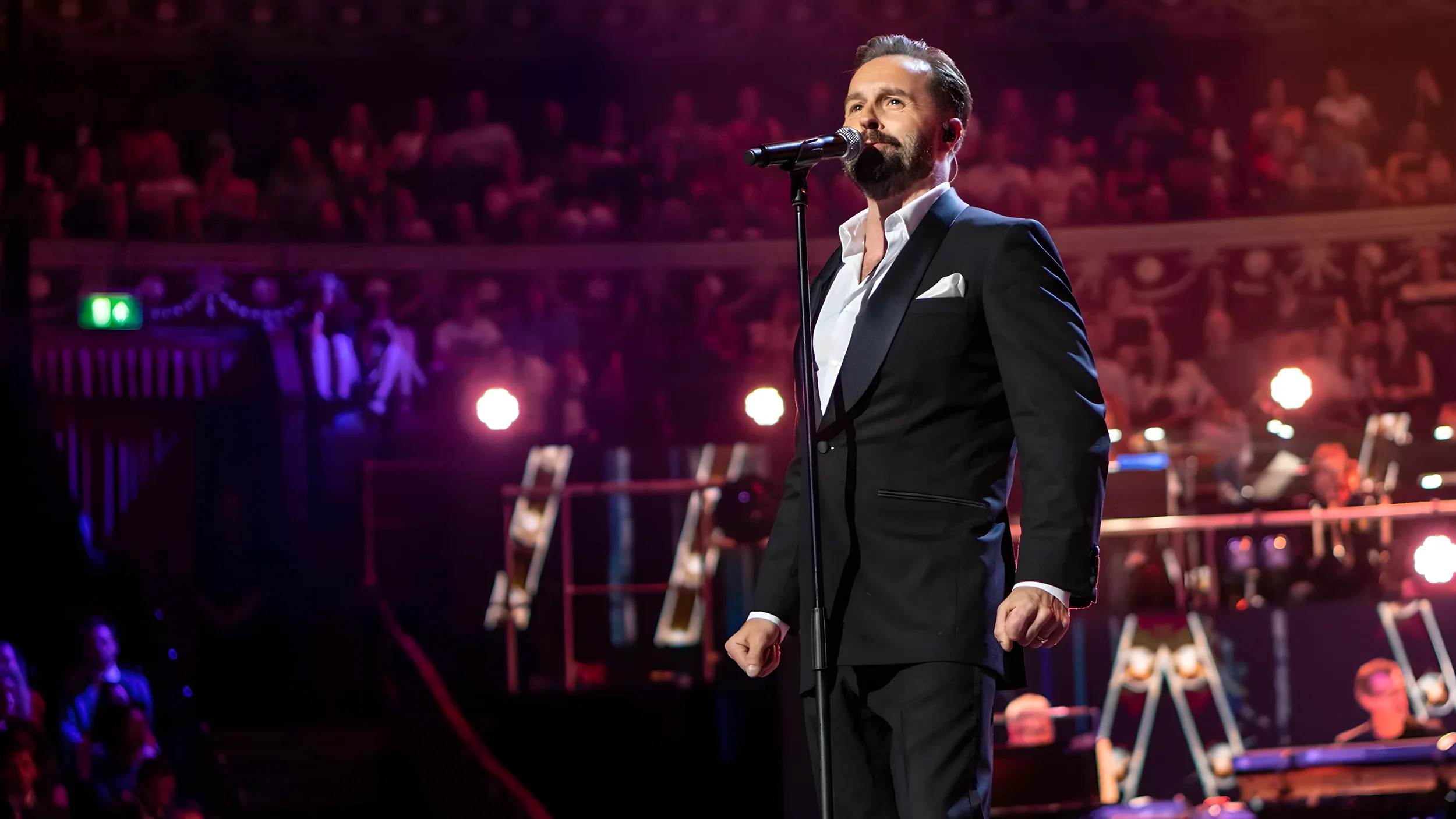 A man, dressed in a black tuxedo with a white shirt and pocket square, is standing on stage singing into a microphone, with an orchestra and audience in the background.