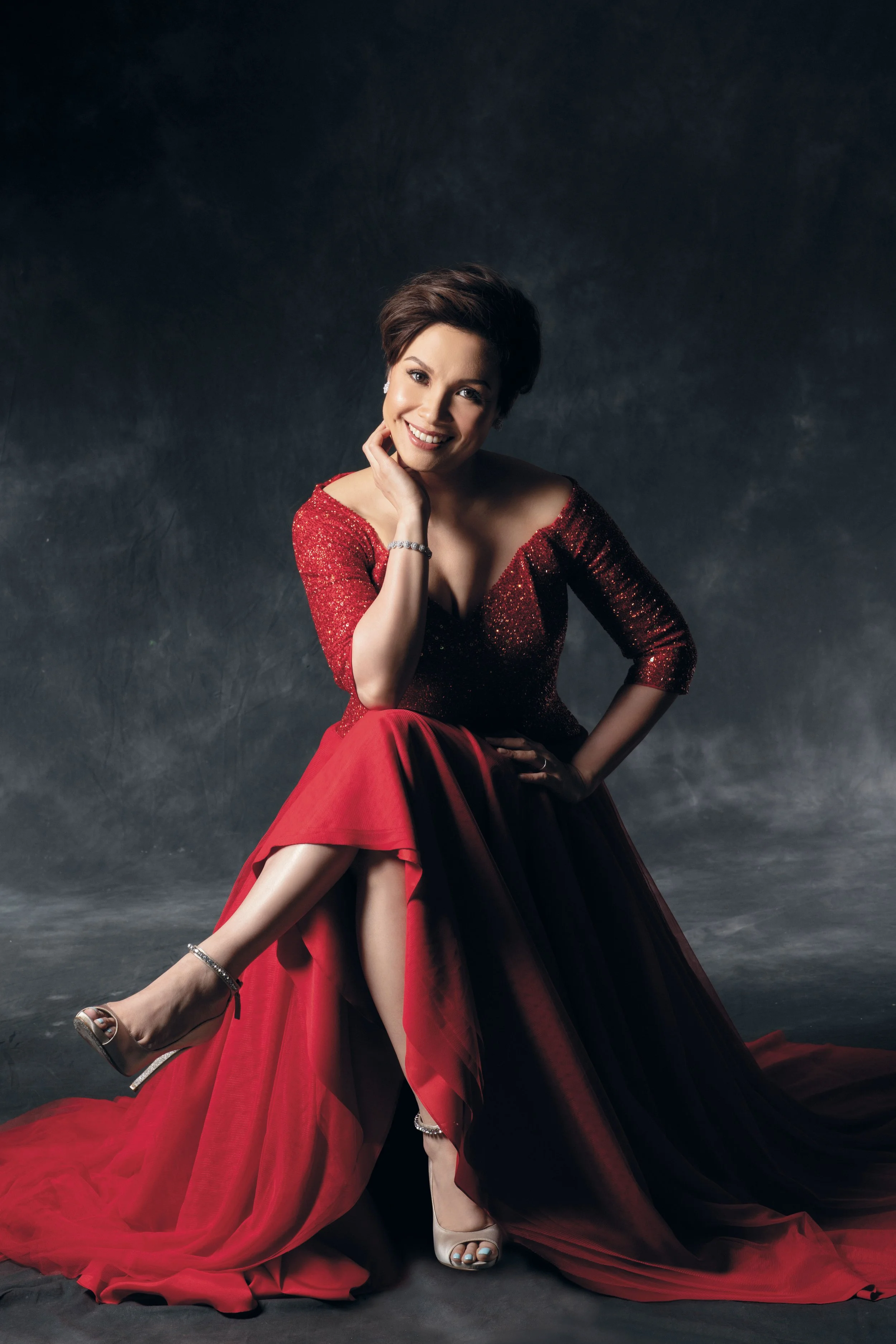 A woman in a red sequined top and long red skirt sitting with one leg crossed over the other, smiling, against a dark background. She is wearing silver jewelry and high heels.