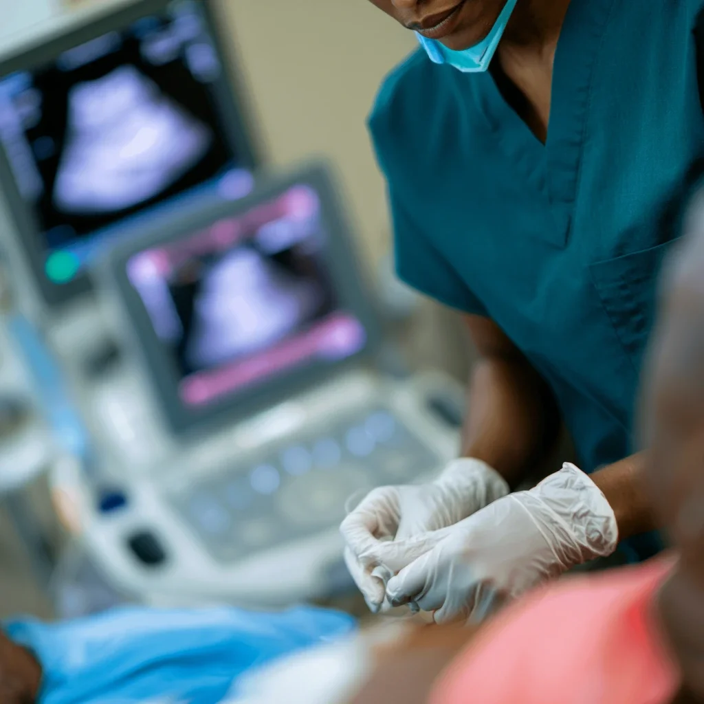 Medical professional wearing gloves, scrubs, and a face mask, examining a patient in a clinical setting, with an ultrasound machine in the background.