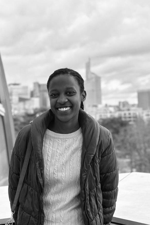 A smiling woman with braided hair wearing a white sweater and puffy jacket, standing in front of a cityscape with tall buildings, during cloudy weather.