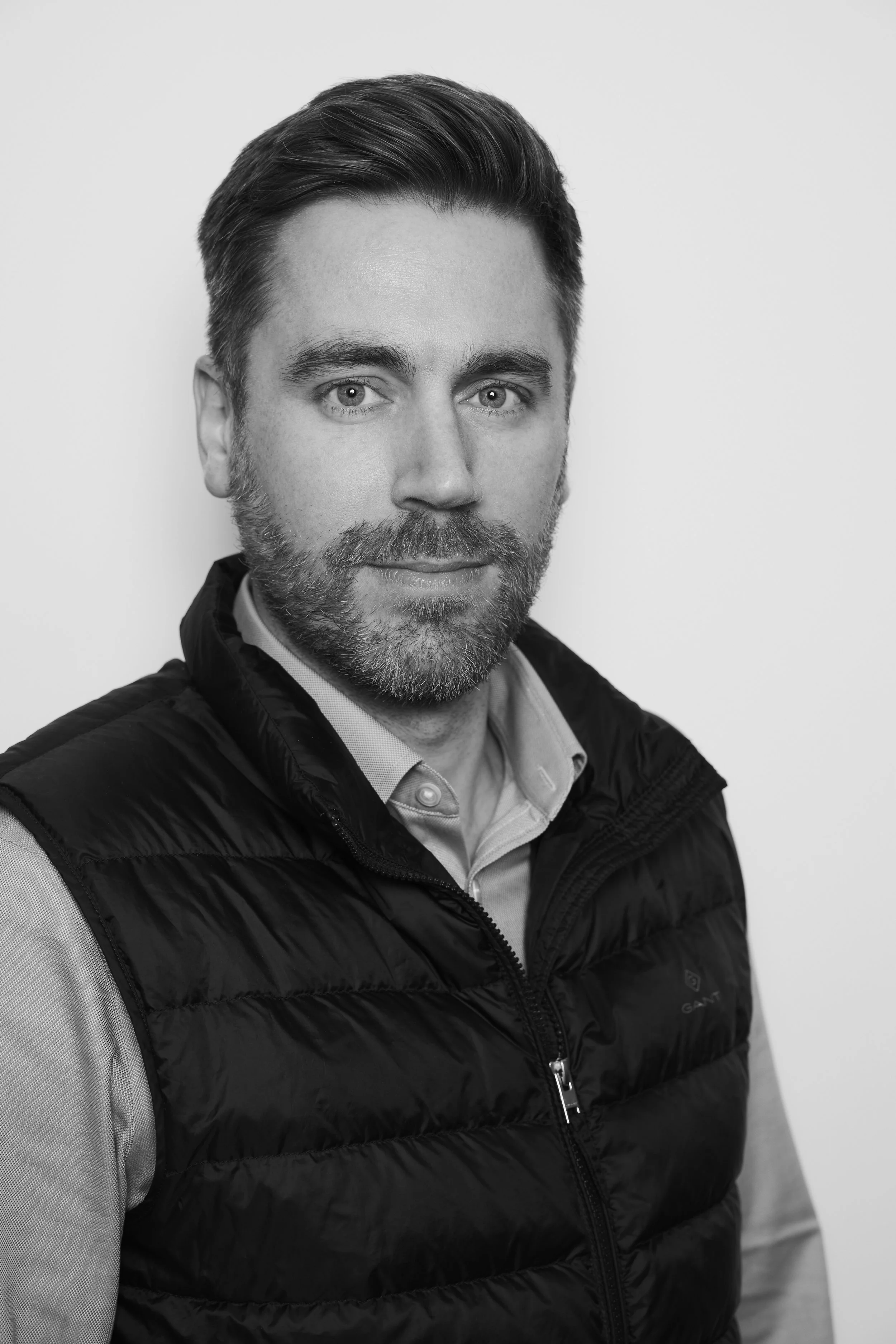 Black and white portrait of a man with a beard and styled hair, wearing a light-colored shirt and a black quilted vest, looking directly at the camera against a plain light background.