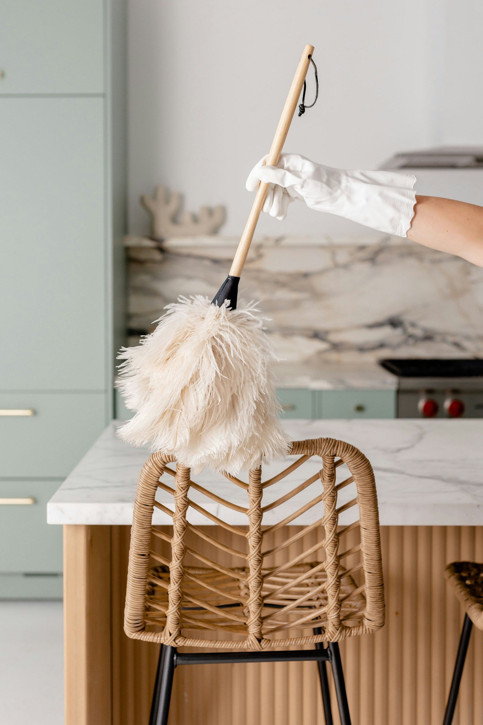 A wicker chair with a black metal frame is positioned next to a white marble kitchen island. A bunch of fluffy lint-free cloths is draped over the back of the chair. A person's right arm, wearing a white rubber glove, is holding a dust mop with a wooden handle, cleaning the cloths. The background features a light green kitchen cabinet, a marble backsplash, and a stove with red knobs.