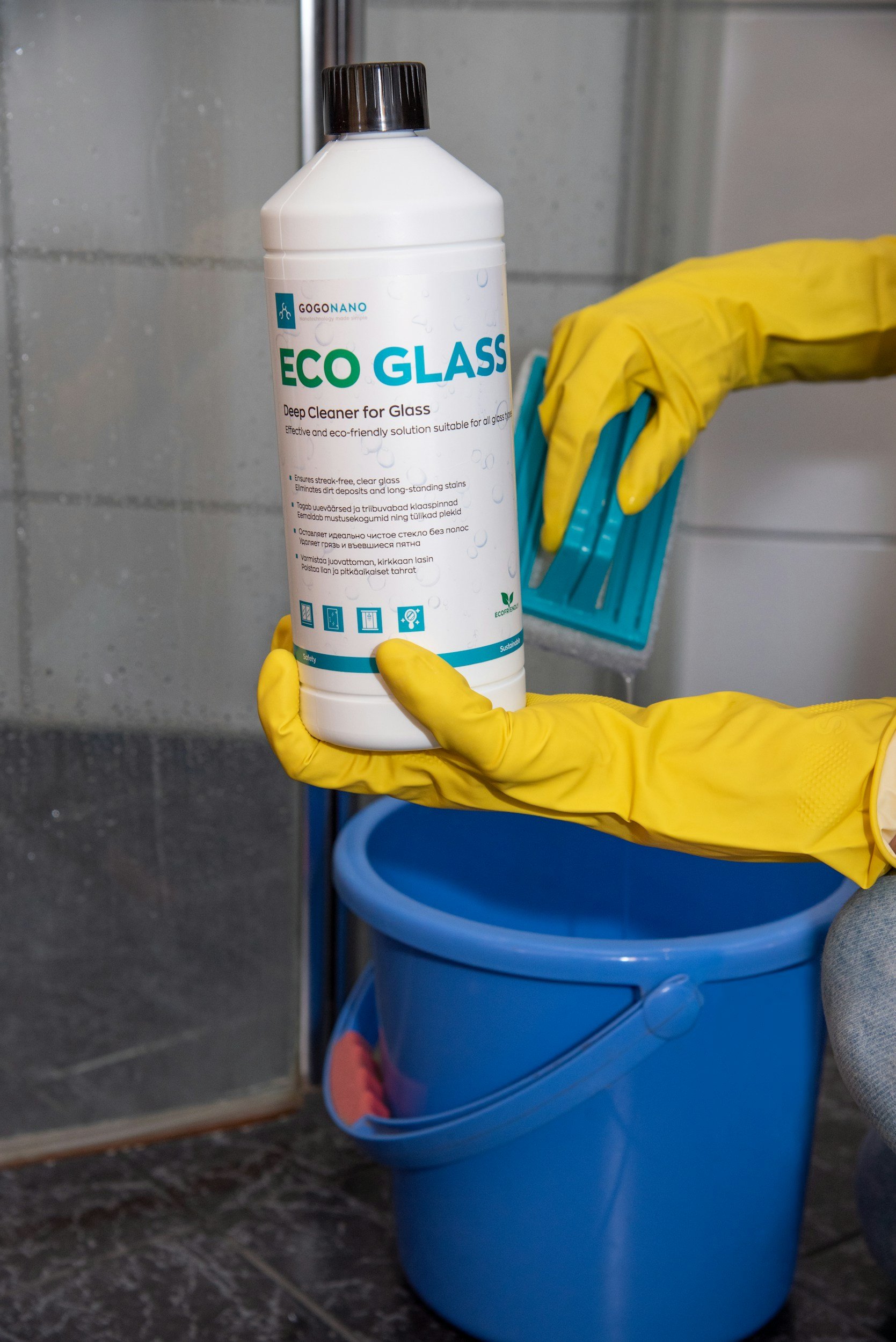 A person wearing yellow rubber gloves holding a white bottle of Eco Glass deep cleaner near a blue bin with a pink cloth inside. The background shows a metal sink and a gray tiled wall.