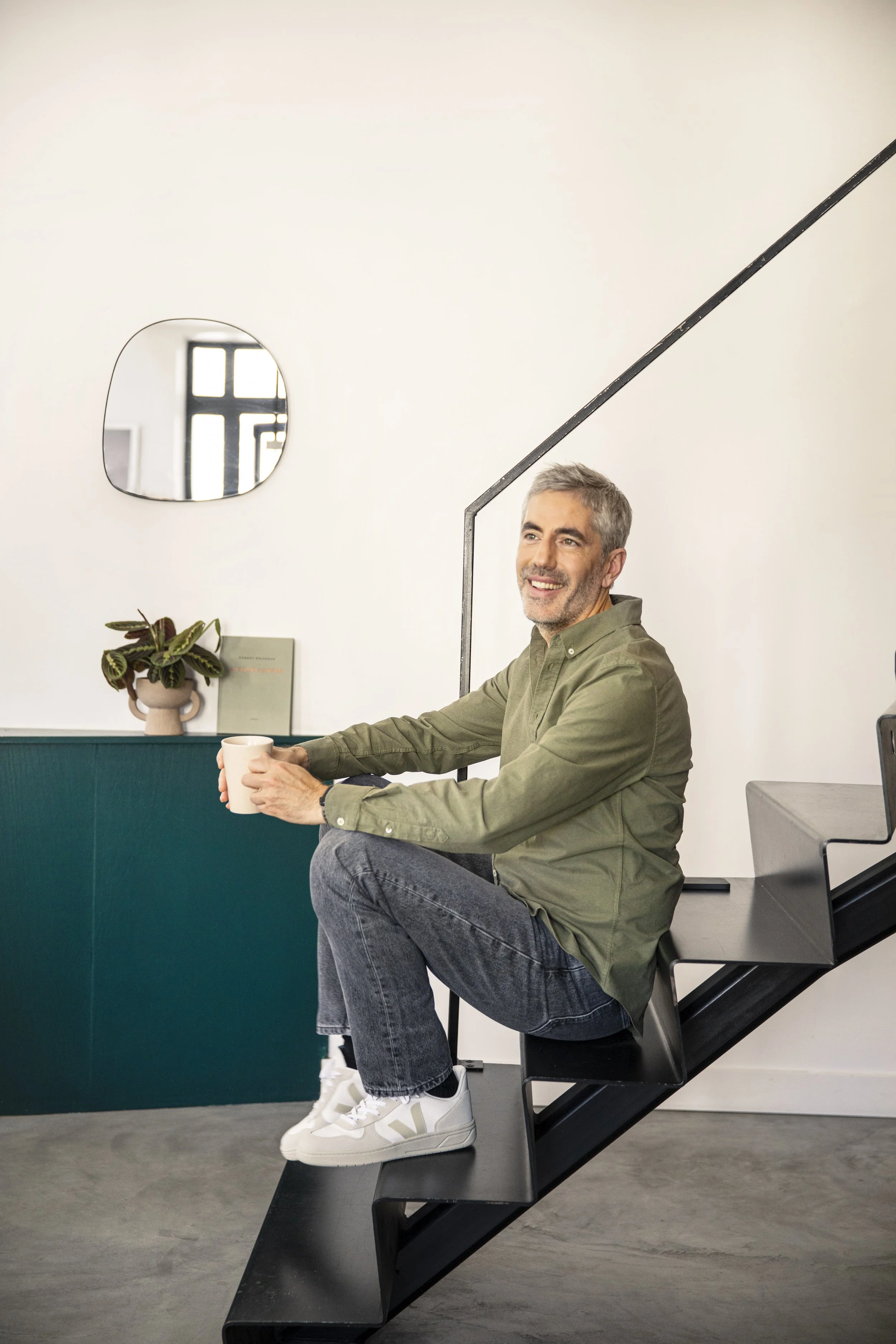 Un homme assis sur une banquette noire à côté d'une staircase noire, tenant une tasse blanche. Décoration minimaliste avec une plante dans un pot beige, un livre vert, un miroir et une fenêtre derrière lui, dans un intérieur moderne.