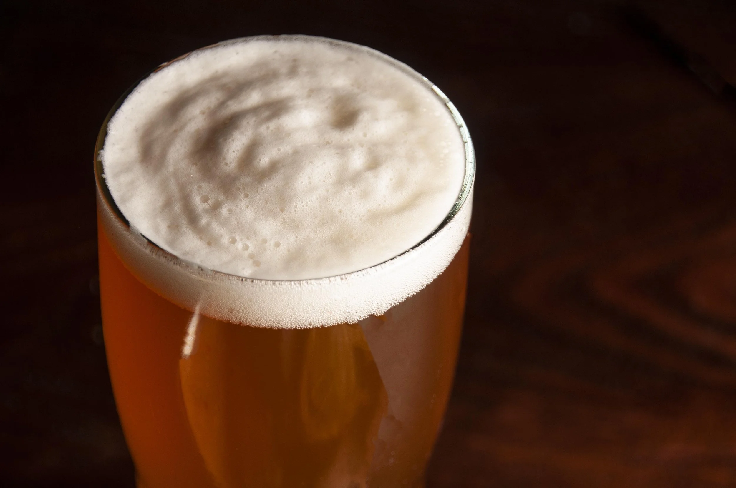 Close-up of a glass of beer with a frothy head on a dark surface.