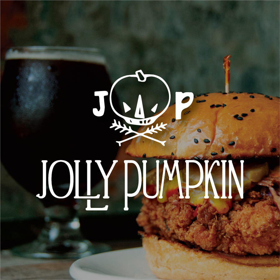 A close-up of a fried chicken sandwich with black sesame seeds on a bun and a glass of dark beer in the background. Jolly Pumpkin, Ann Arbor, Michigan. Traverse City, Michigan. East Lansing Michigan.