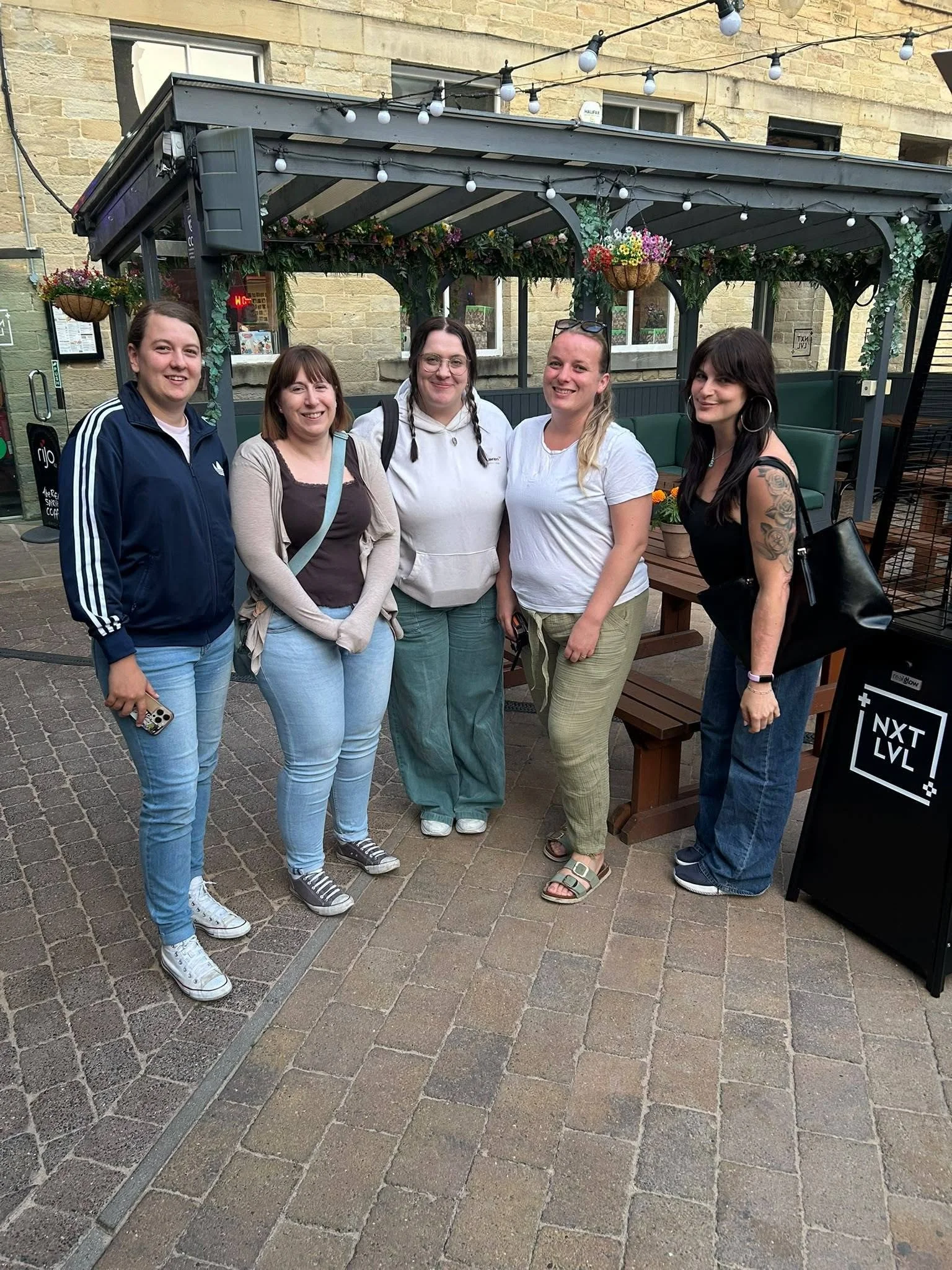 Group of five women standing together outdoors on brick pavement in front of a building with a covered patio and hanging flower baskets, smiling at the camera.