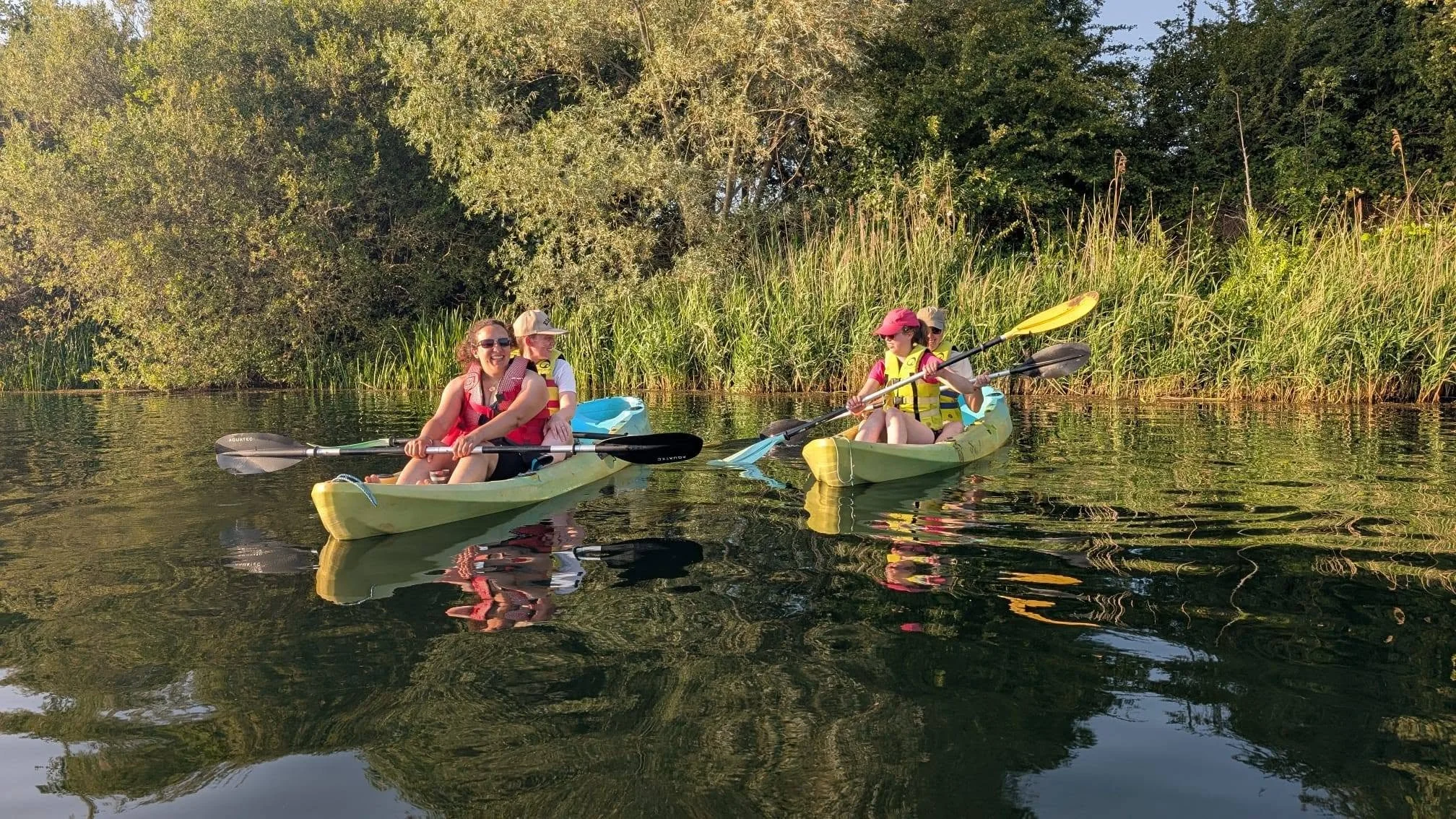 Two kayaks with groups of people enjoying a paddle on a calm river or lake surrounded by lush greenery and trees.
