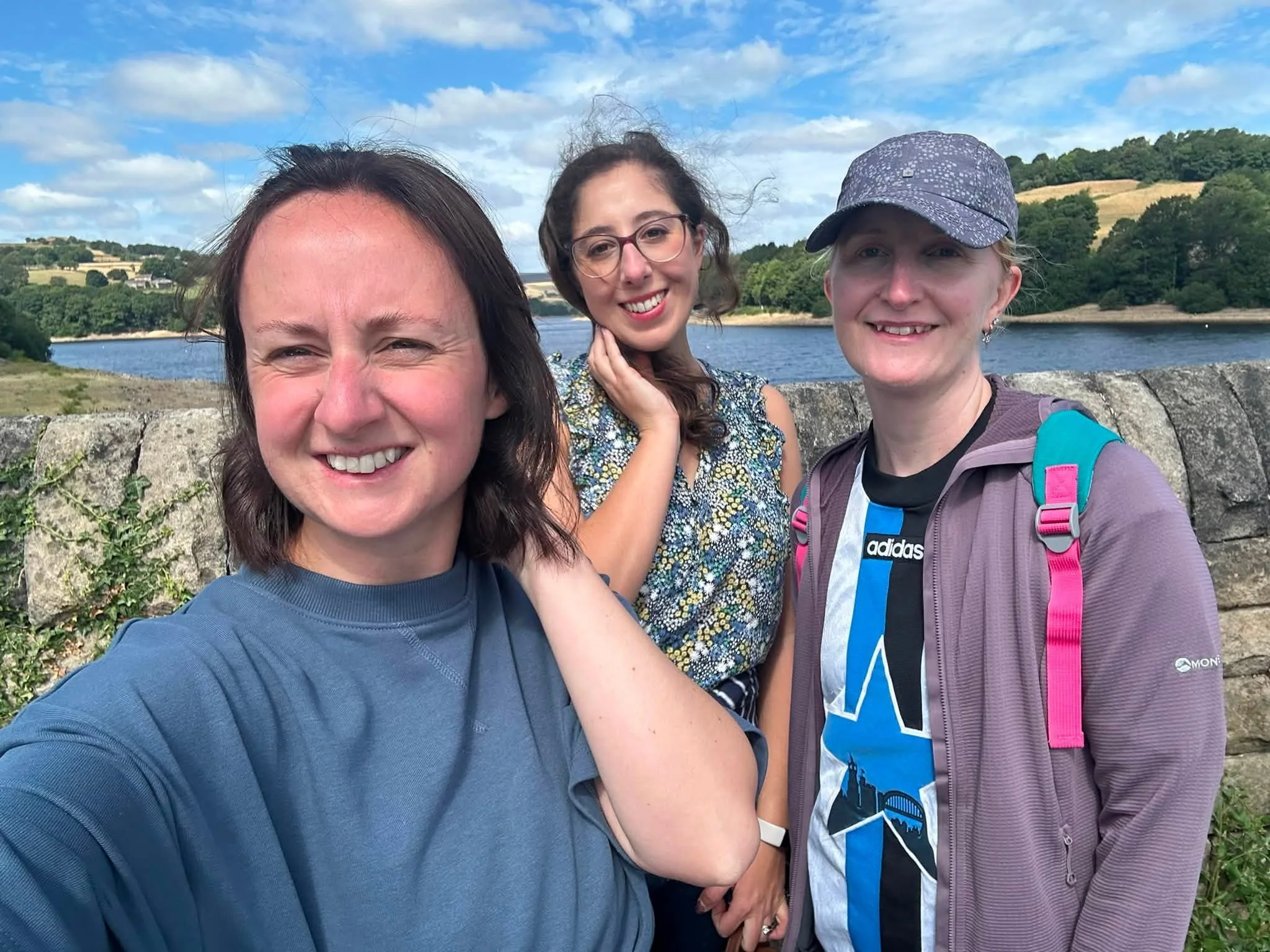 Three women smiling and posing for a selfie outdoors near water with hills and trees in the background. One woman is wearing a blue shirt, another is in glasses with a floral top, and the third is wearing a purple cap and a backpack.