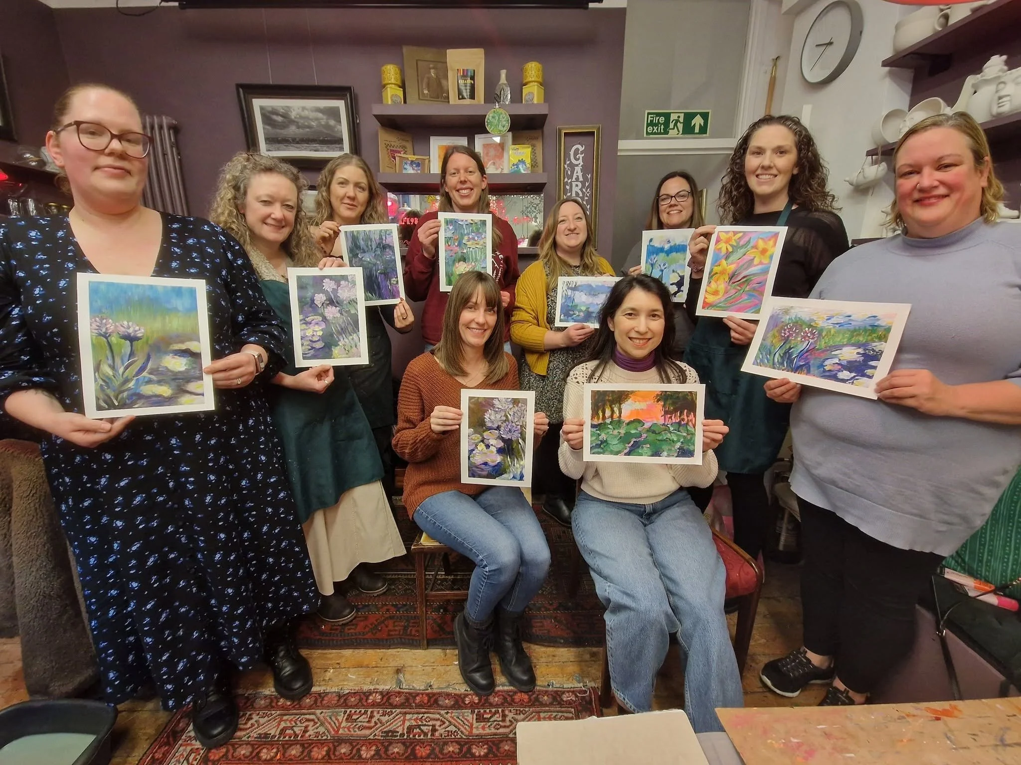 A group of nine women in an art classroom or studio, each holding a colorful watercolor painting of flowers or nature scenes, smiling at the camera.