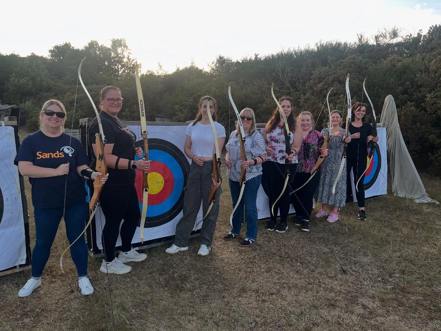 Group of women holding bows and arrows at an outdoor archery range with targets in the background.