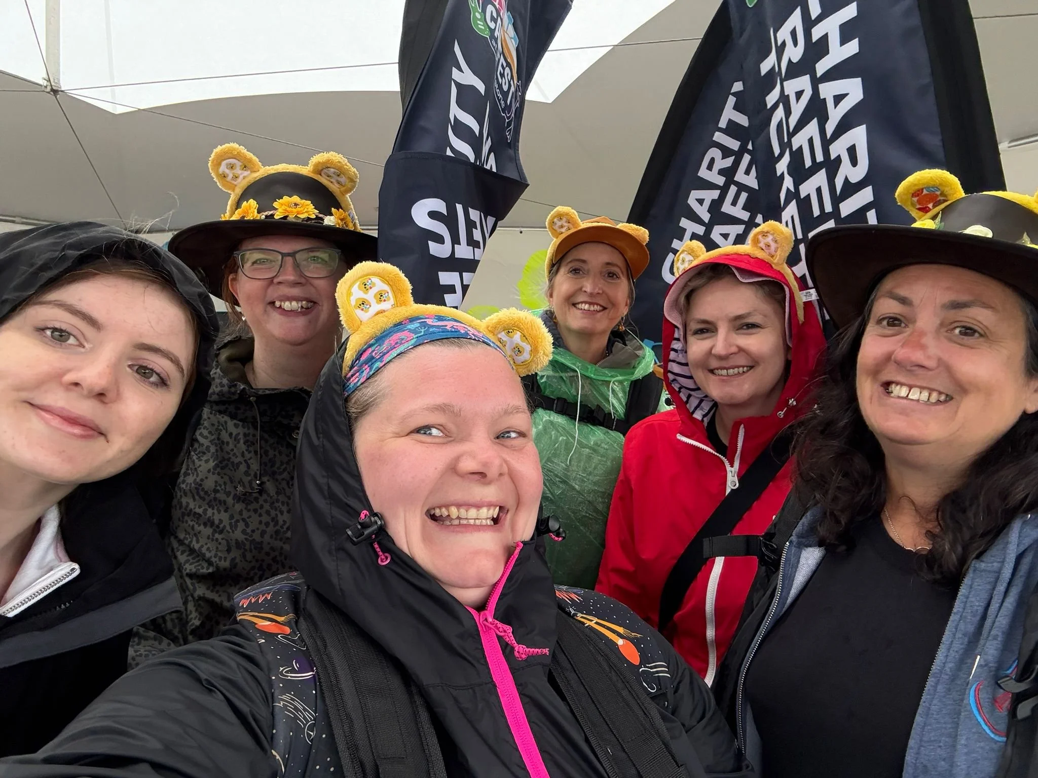 A group of smiling women wearing colorful hats with bear ears and outdoor jackets, posing together under a white tent. Two black banners with white and orange text are visible in the background.