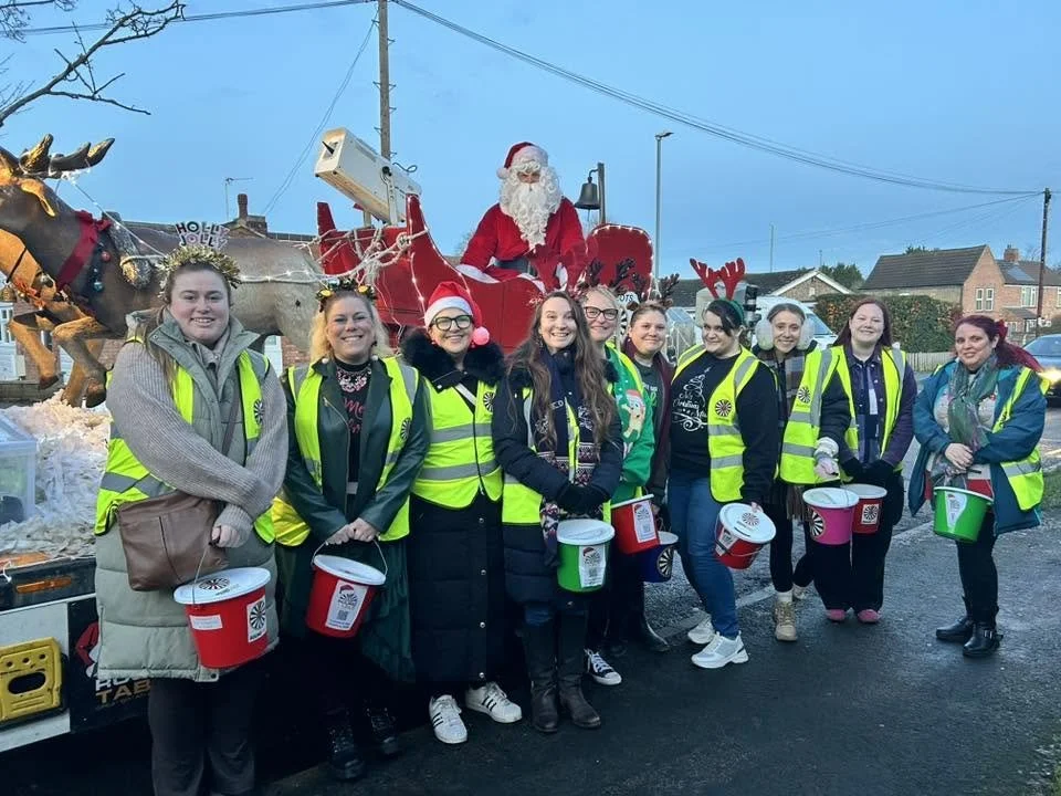 Group of women wearing yellow safety vests and Christmas accessories, standing in front of a Christmas float with Santa Claus and reindeer.