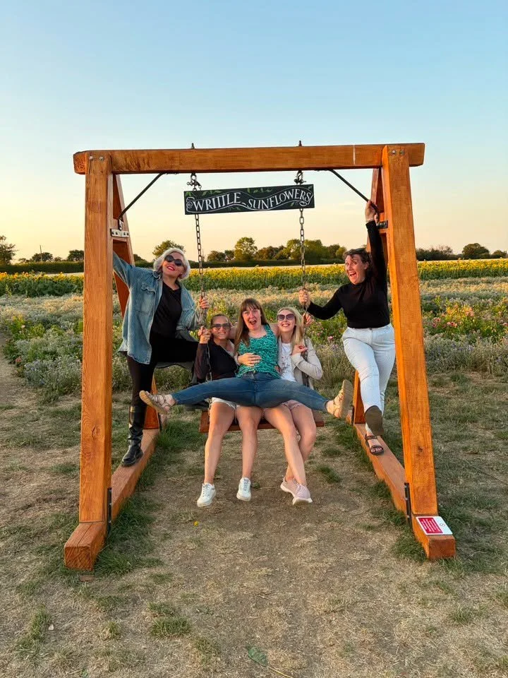 Five women are sitting and standing on a large wooden swing in a sunflower field during sunset. They are smiling and enjoying themselves, with some holding drinks and one woman doing a split on the swing.